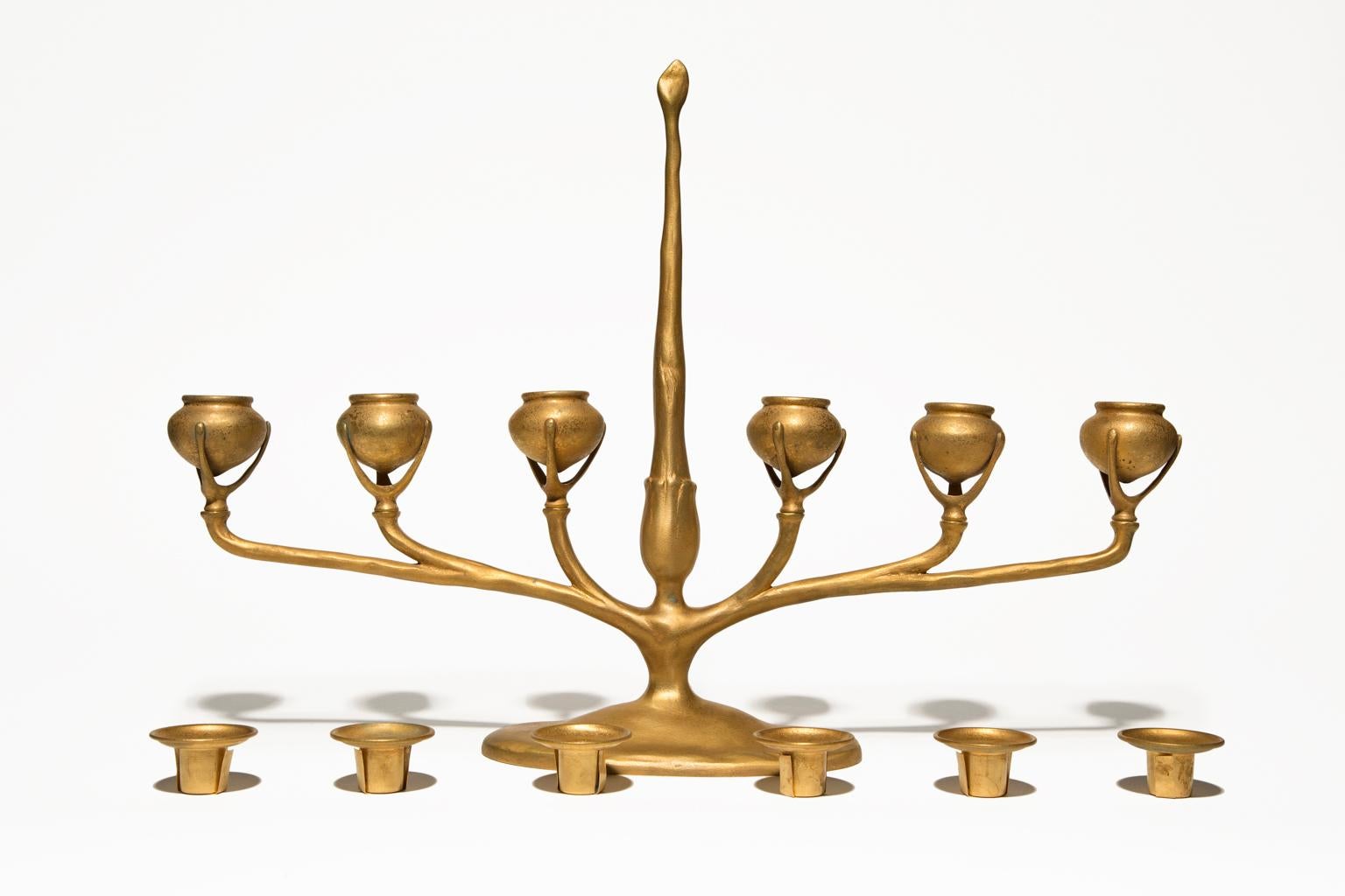 This exquisite antique is a Tiffany Studios gilted bronze candelabra from the early 20th century. It features six arms for candle cups and a center piece that rises above it like a spire. The candle cups are urn form cups and are outfitted with
