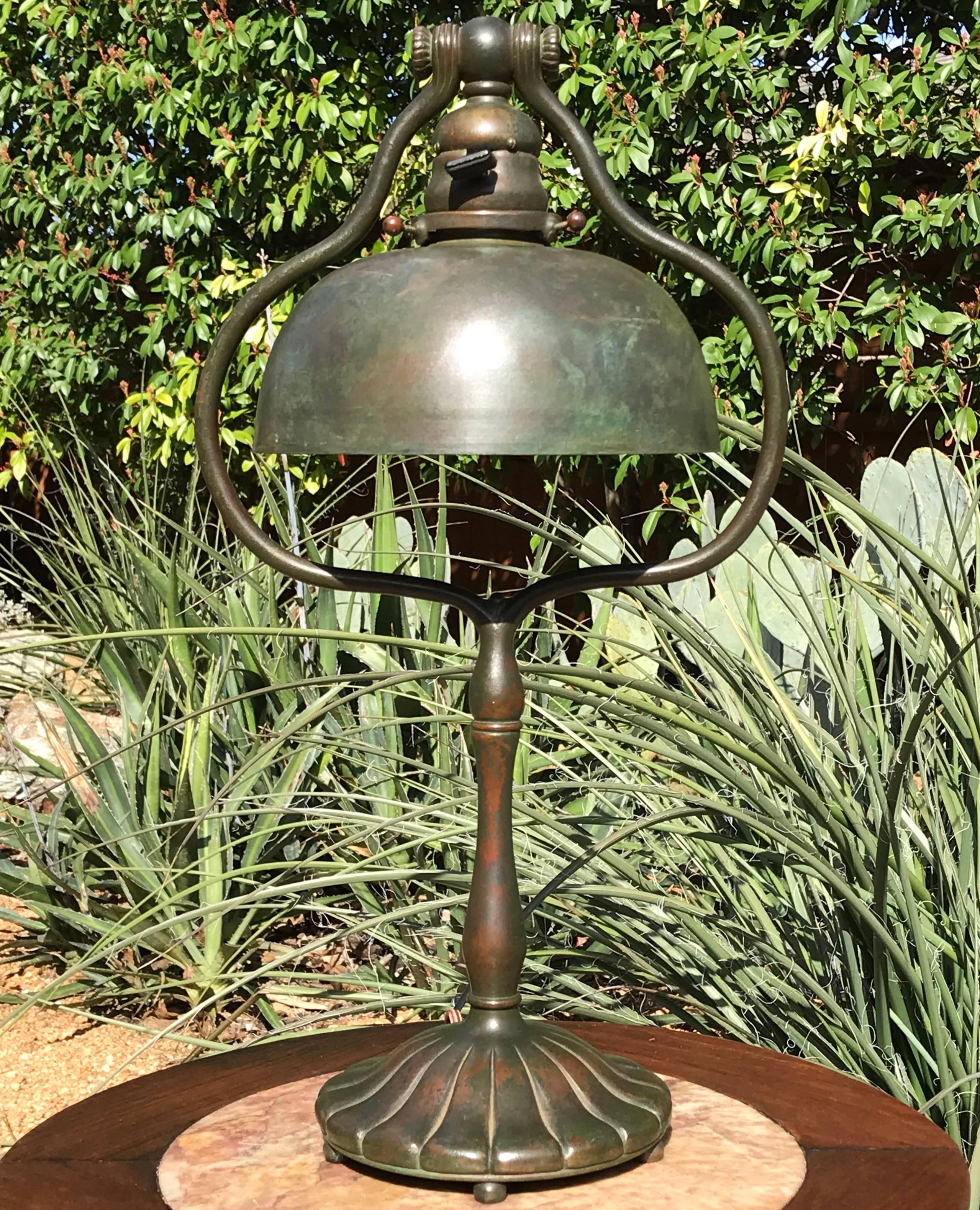 A tall elegant Tiffany Studios bronze desk or table lamp with original patinated bronze shade. The green and reddish brown base with scrolled bulbous harp form and terminating on a circular fluted base with five ball feet. Shade has a colorful