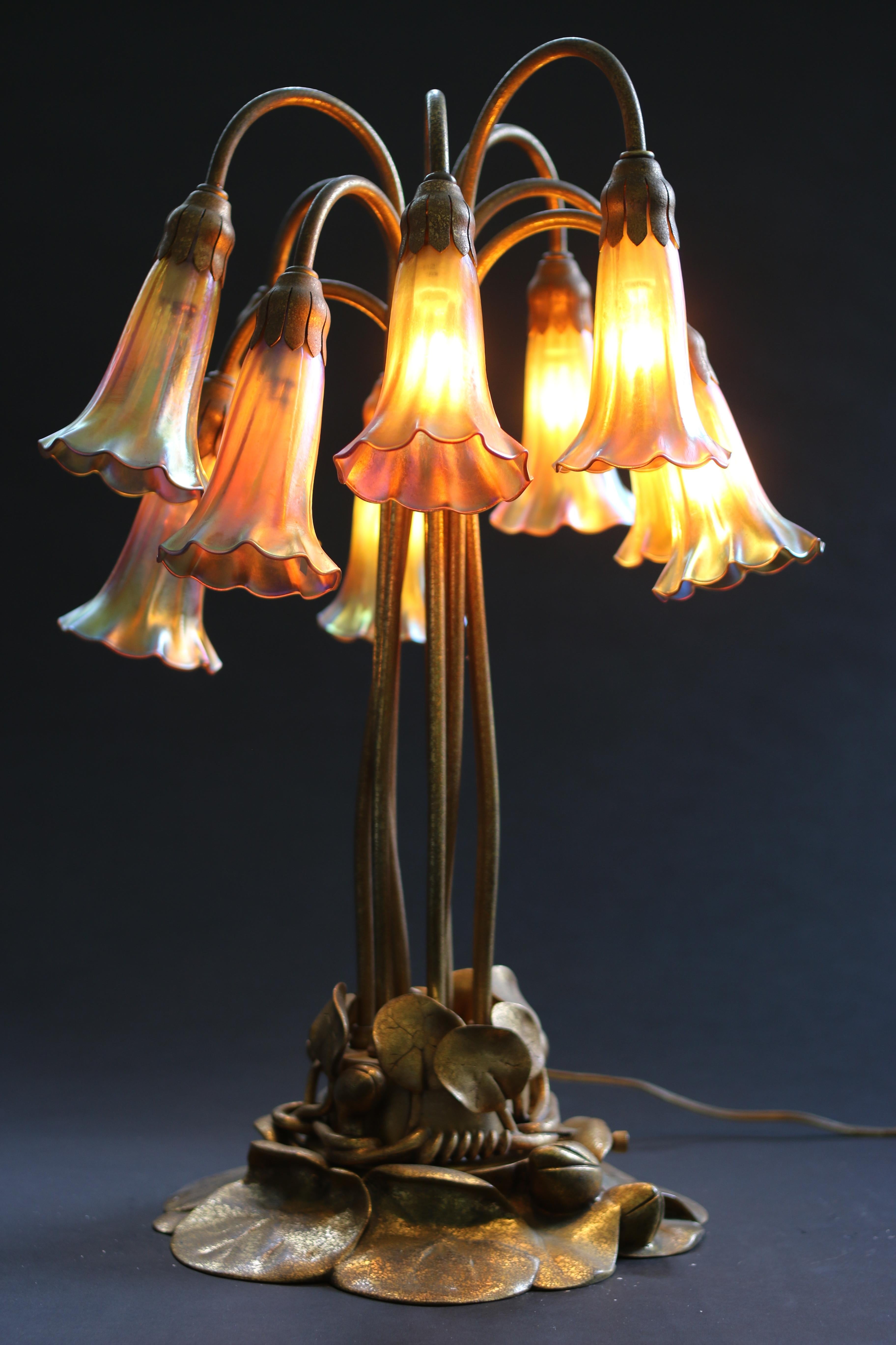 Here is a Tiffany Studios gold 10-light lily lamp with the a lily pad base and elegant stems that terminate in 10 authentic Tiffany favrile iridescent lily shades. The shades are well matched in both color and shape. The base is in excellent shape