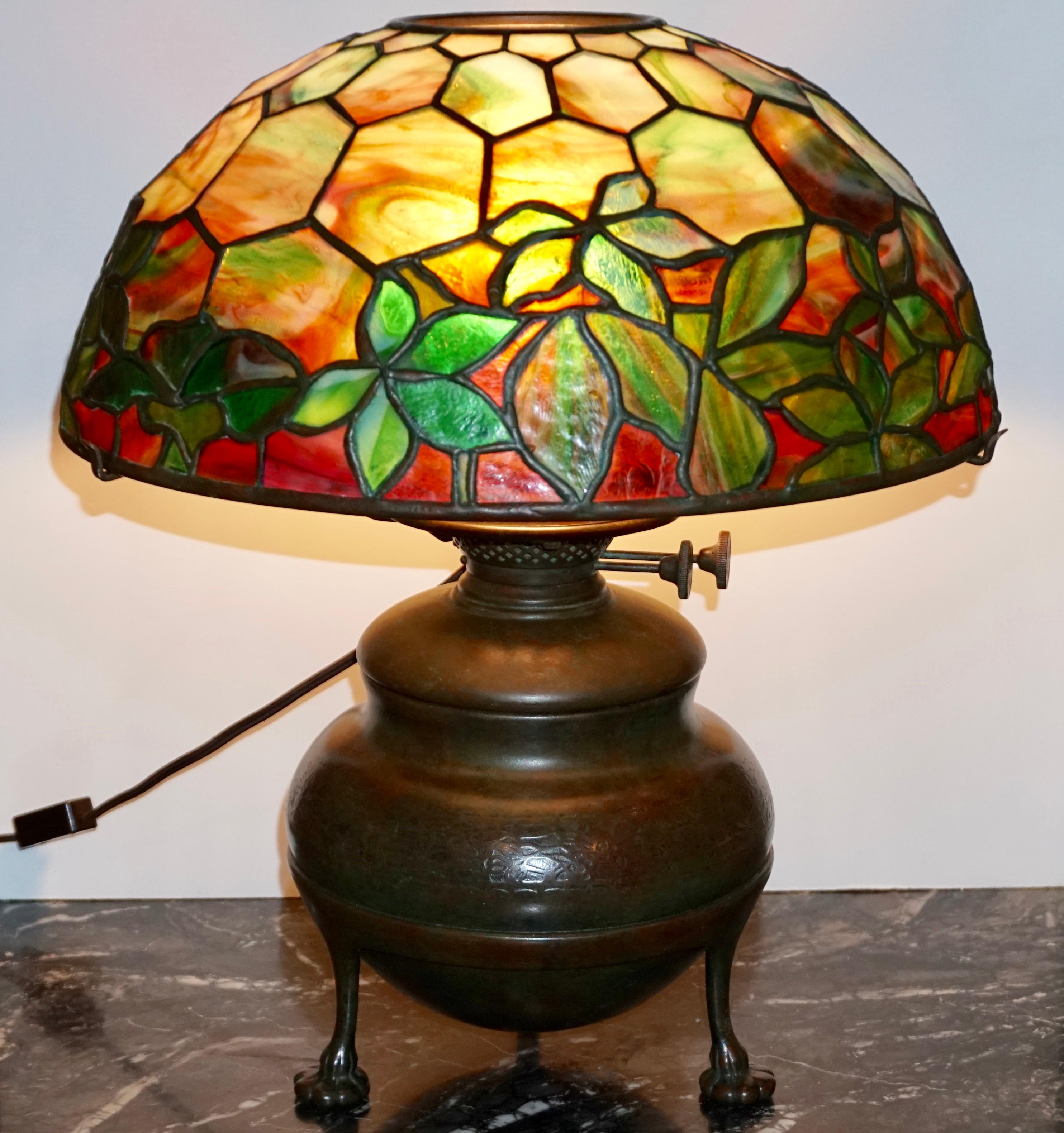 A wonderful Art Nouveau Tiffany studios stained glass Woodbine shade with reds, greens, yellows oranges and combinations of all of the above. Shade sits on a bronze acorn three foot electrified oil lamp. Base has a wonderful raised design around the
