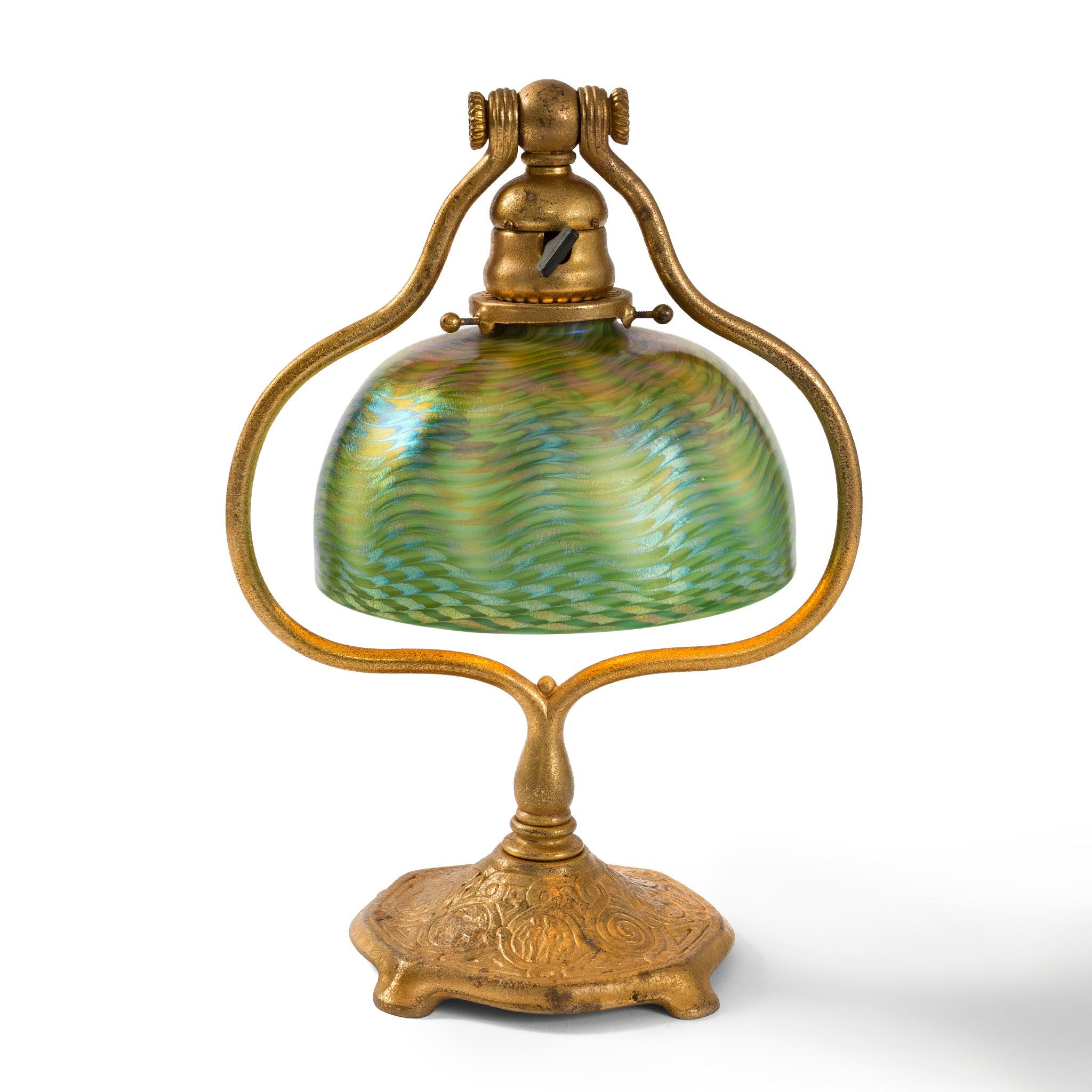 Dating from 1906-1920, this Tiffany studios desk lamp is composed of Damascene favrile glass and a gilt bronze Zodiac base. The green domed shade with golden brown accents exhibits a wave pattern, with subtle iridescent touches, and is suspended