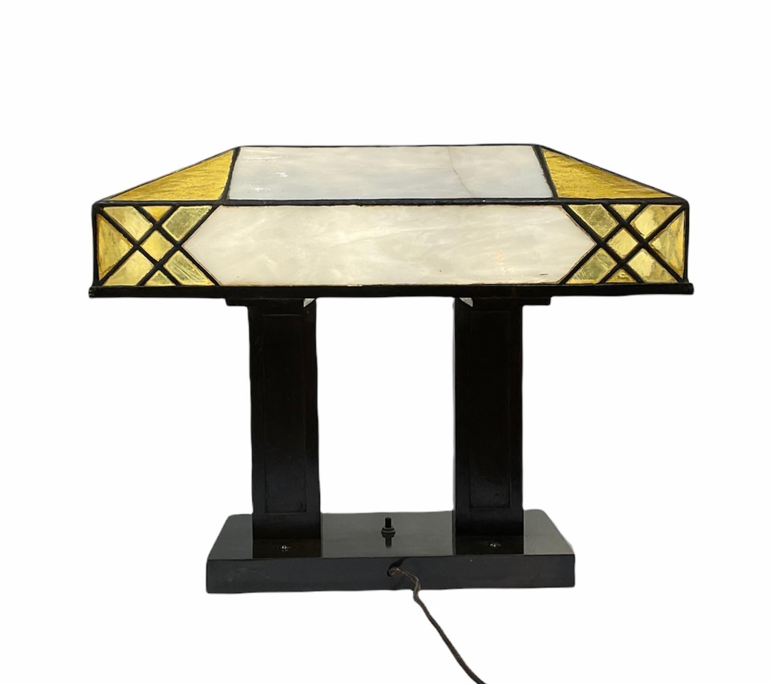 This is a Tiffany style Art Deco stained glass rectangular desk/table lamp. It features a shade made of a mosaic of topaz yellow and white color geometric pieces of stained glass mounted in wrought iron. It is supported by two standing wide metal