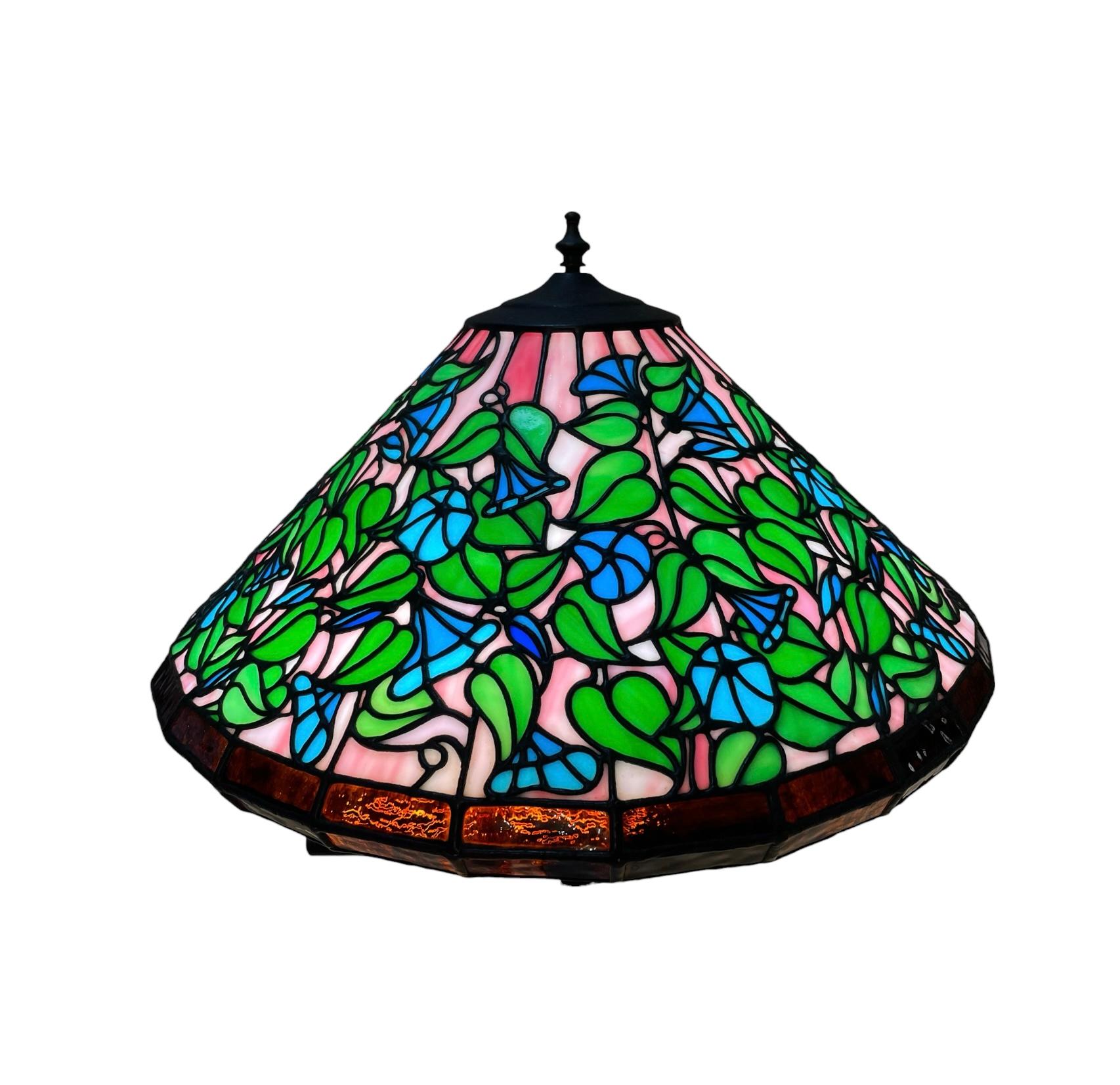 Il s'agit d'une lampe de table en bronze et en verre teinté. Elle représente une lampe à pied en bronze ornée au centre d'une guirlande repoussée de cupidons jouant d'instruments de musique, certains d'entre eux chevauchant un bélier. Le haut de la