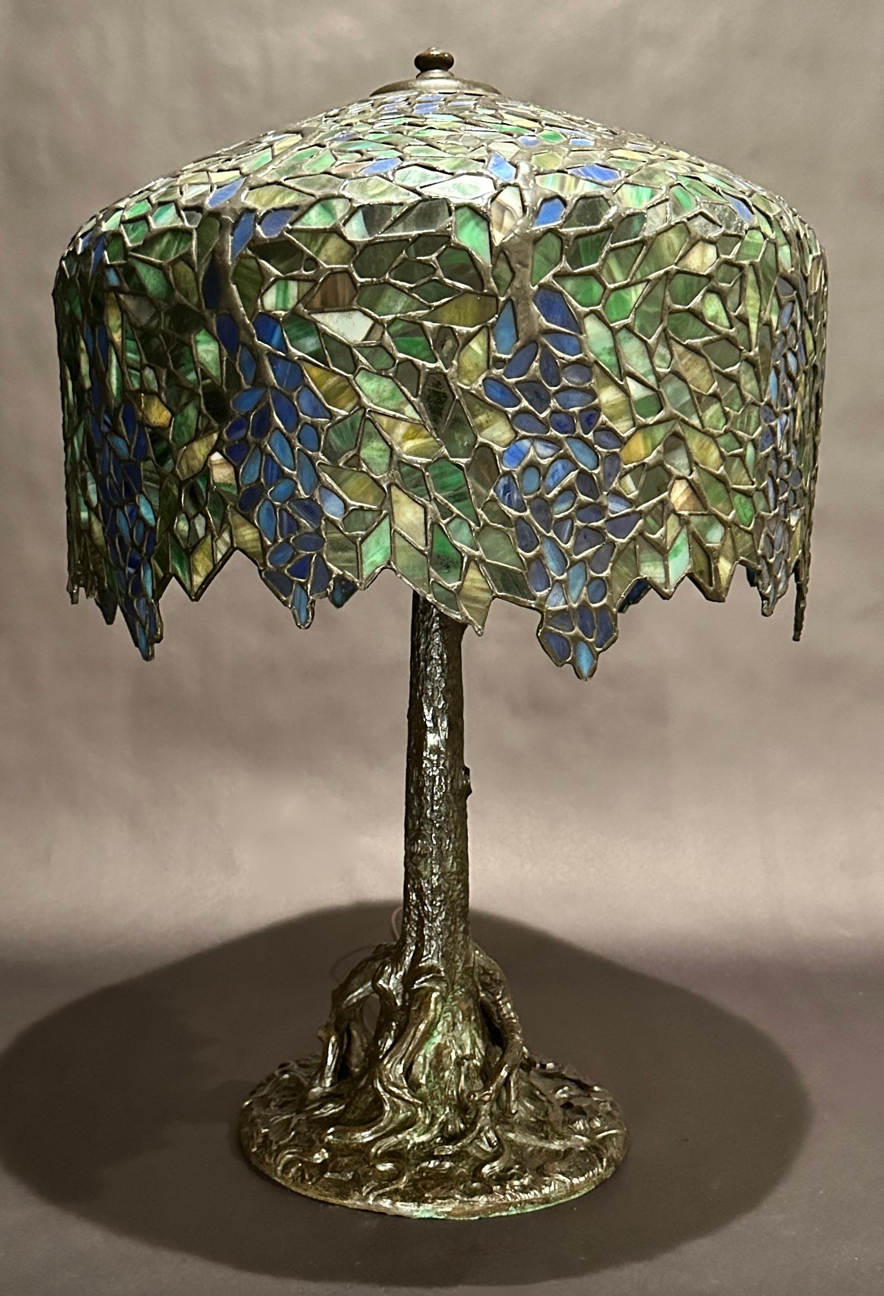 Fine quality beautiful bronze tree trunk form lamp base with domed leaded glass shade in wisteria pattern. Base marked Handel. Shade featuring varying colors of greens, yellows and blues. Bronze lamp base with beautiful rich brown and green patina.