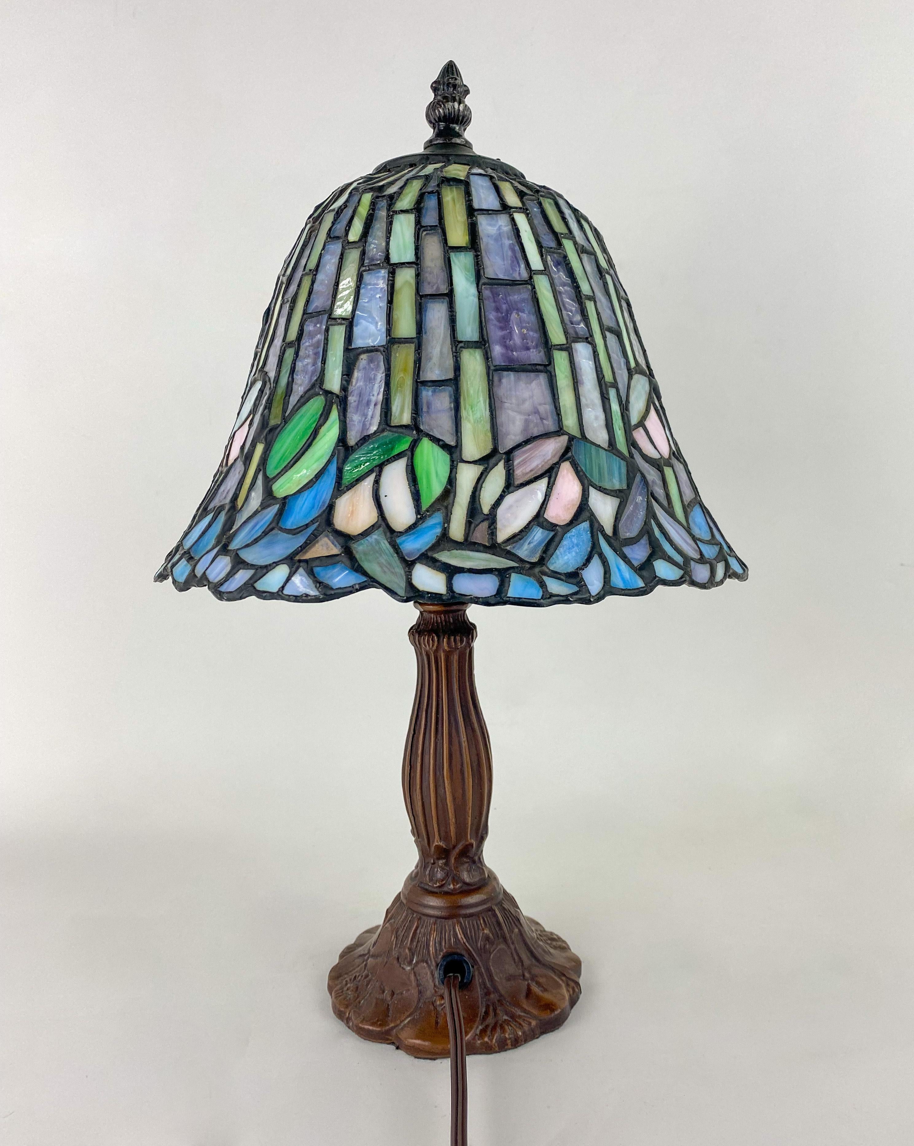 A timeless Tiffany style table lamp. The lamp features multicolor stained glass shade in green, blue, turquoise, purple and pink. The hand cut glass pieces form a beautiful wisteria flower design. The beautiful shade is attached to a patinated