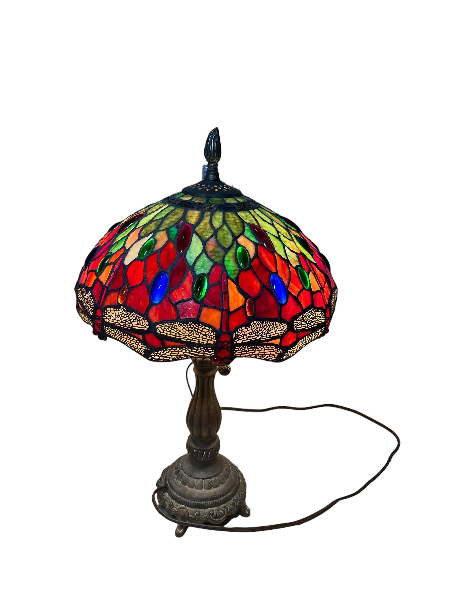 This is a Tiffany Style table lamp. Its shade is shaped as an umbrella. Its stained glass depict different shades of red, orange, yellow and green. These different shapes of geometric glass cuts are joined by a metal. Oval cabochon glasses of