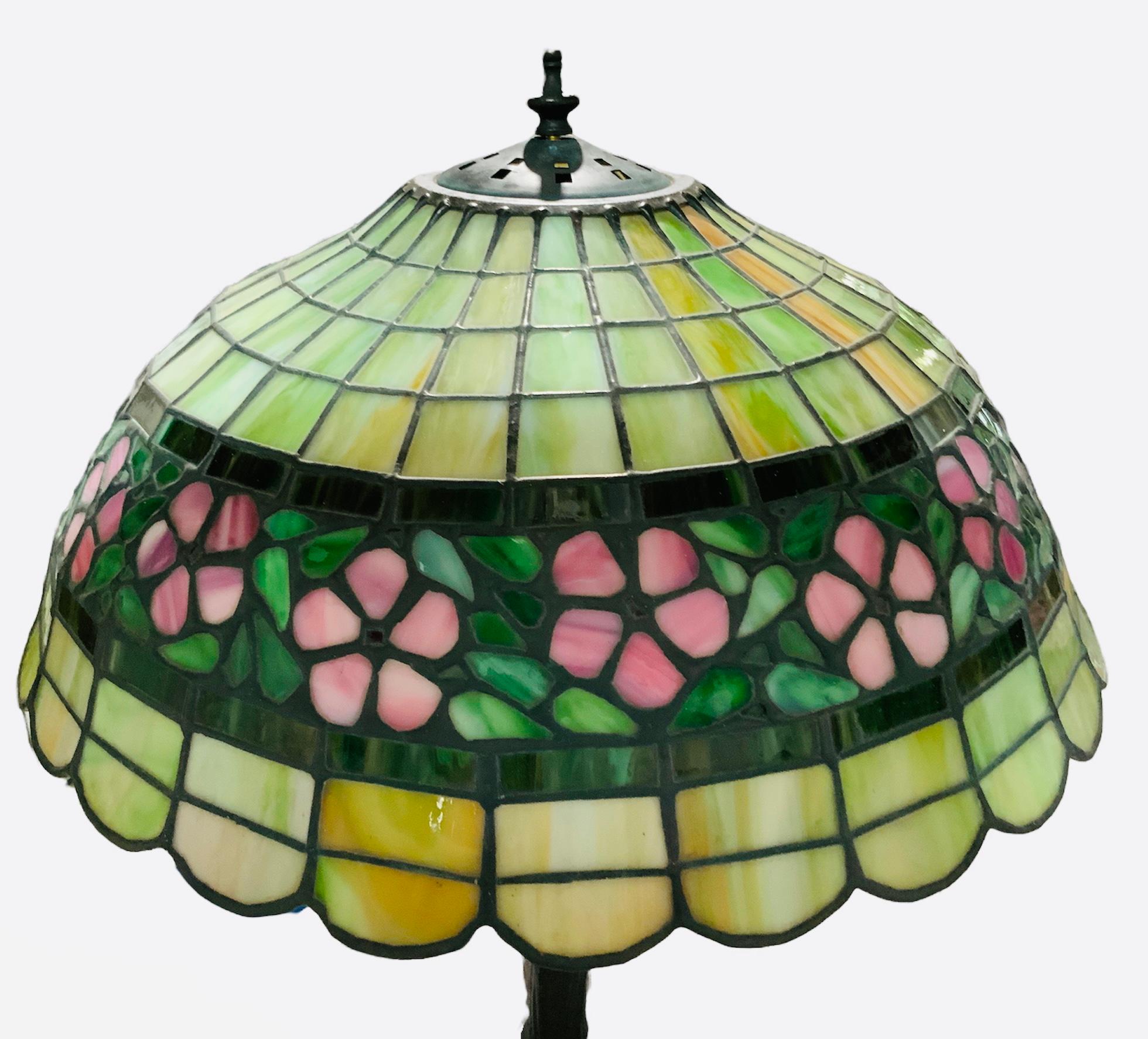 This is a heavy Tiffany & Co. Style table lamp. Its shade is shaped as an umbrella with scalloped border. Its stained glass depict different shades of green, yellow, white and pink. These different shapes of geometric glass cuts are joined by a