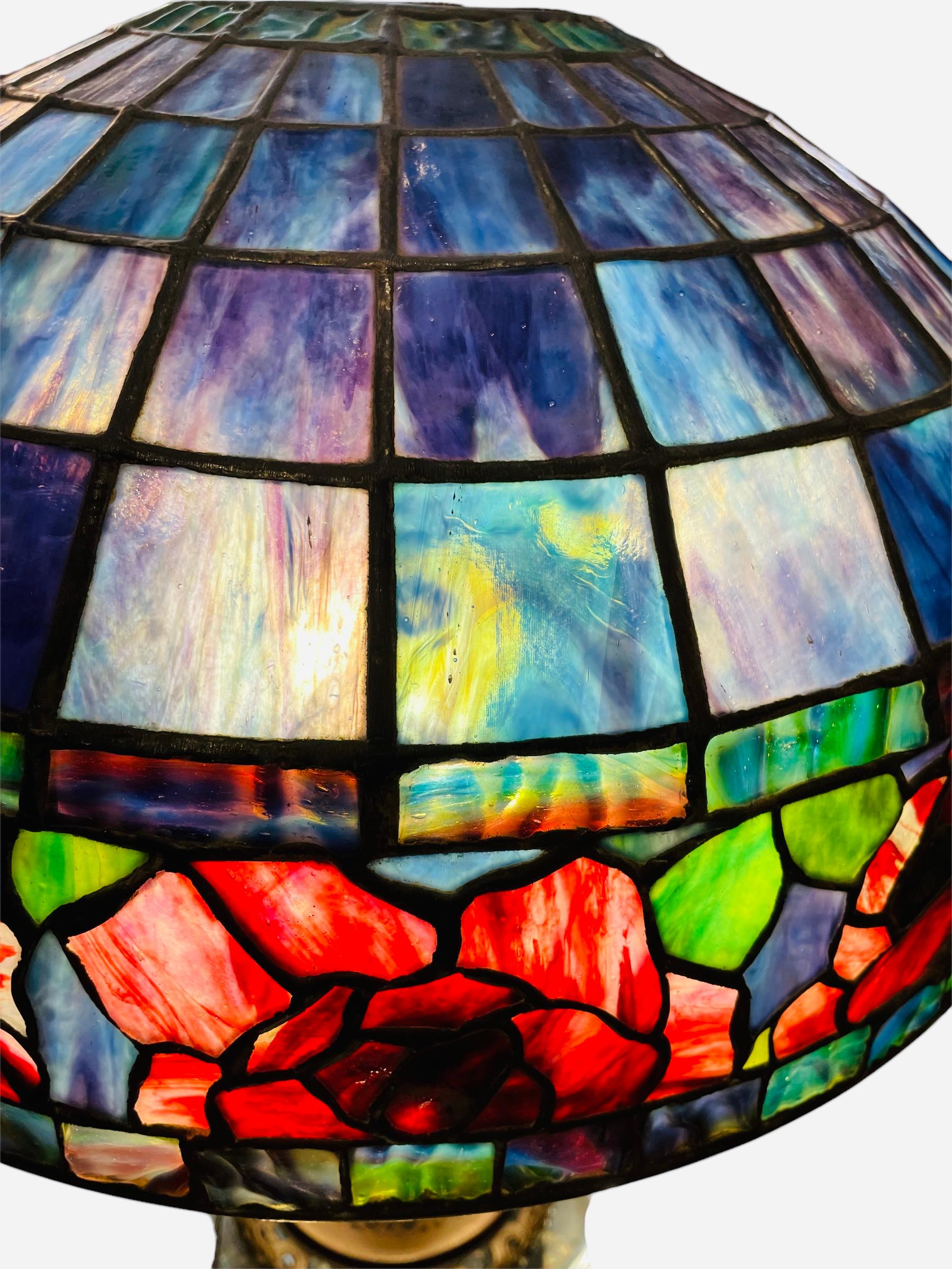 This is a heavy Tiffany Style table lamp. Its shade is shaped as an umbrella. Its stained glass depicts different shades of blue, green, red and purple. These different shapes of geometric glass cuts are joined by a metal. Above the border of the