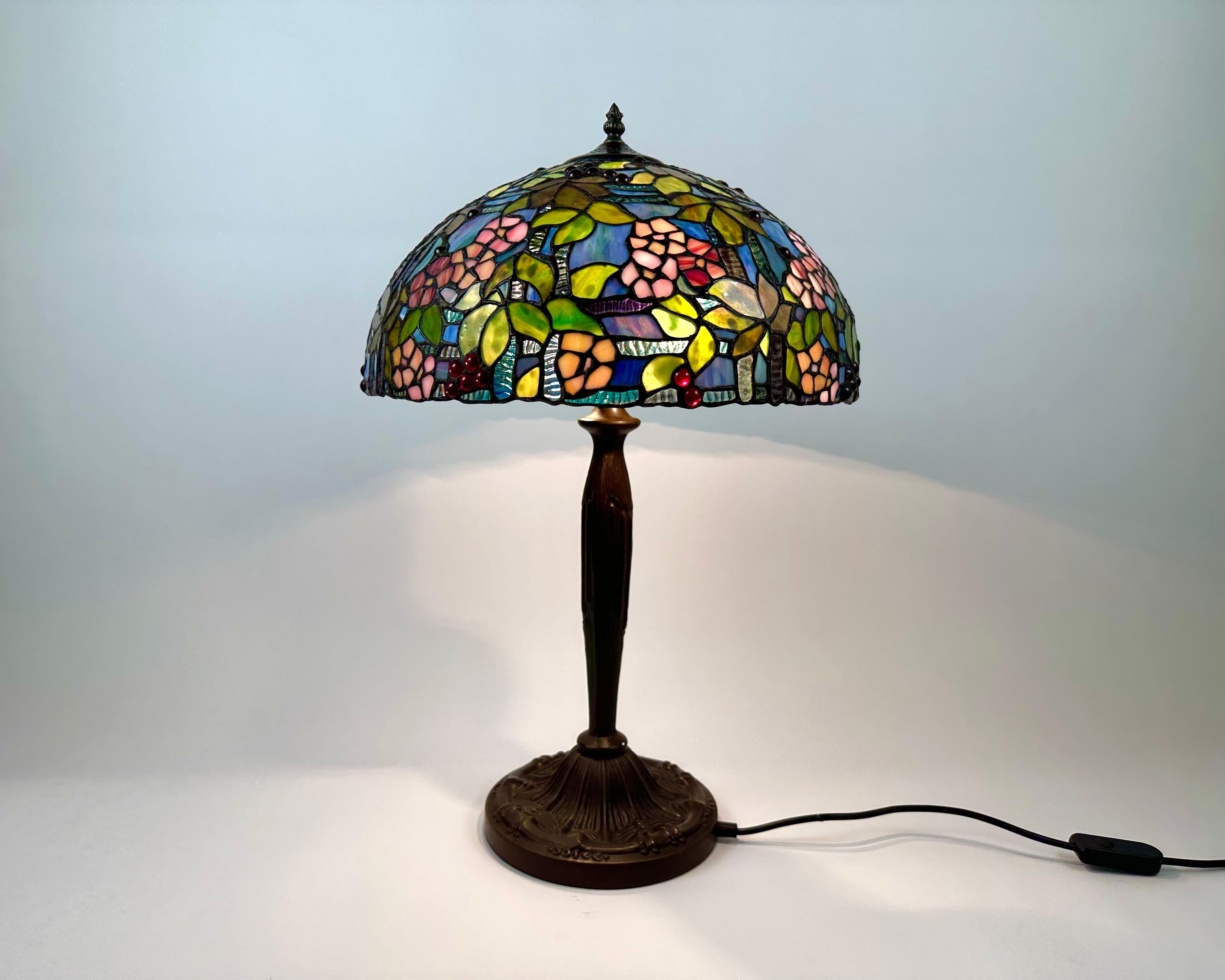 Cette magnifique lampe de table exclusive en verre coloré est fabriquée dans le style classique Tiffany en France, dans les années 1960.

L'abat-jour et la base de cette lampe ont un design unique. La base est en bronze et décorée de belles
