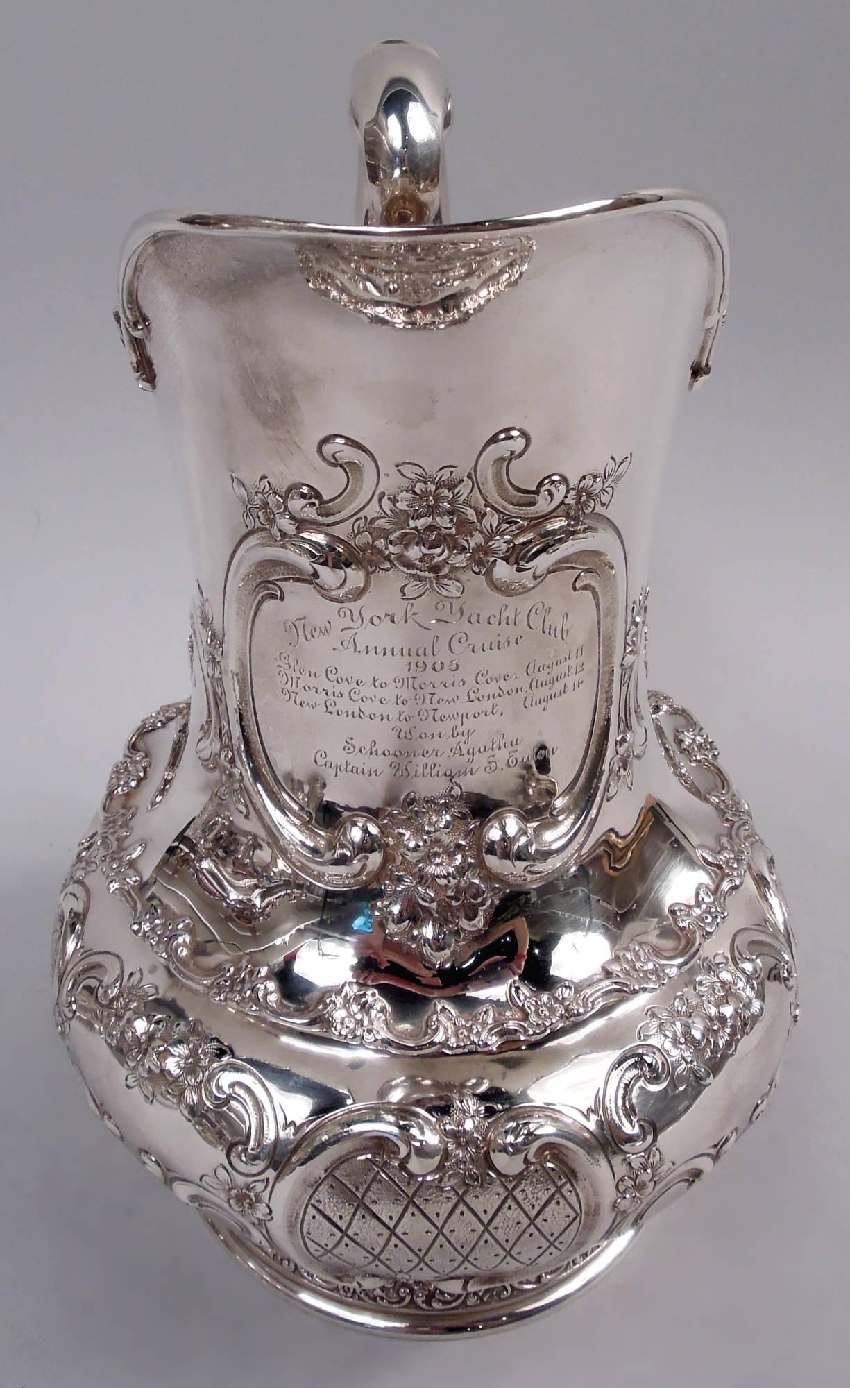 Edwardian Classical sterling silver water pitcher. Helmet mouth and high-looping scroll handle; bellied bowl on domed foot. Chased scroll and flower ornament including 4 vacant frames alternating with 4 diaper frames on bowl.

On neck engraved