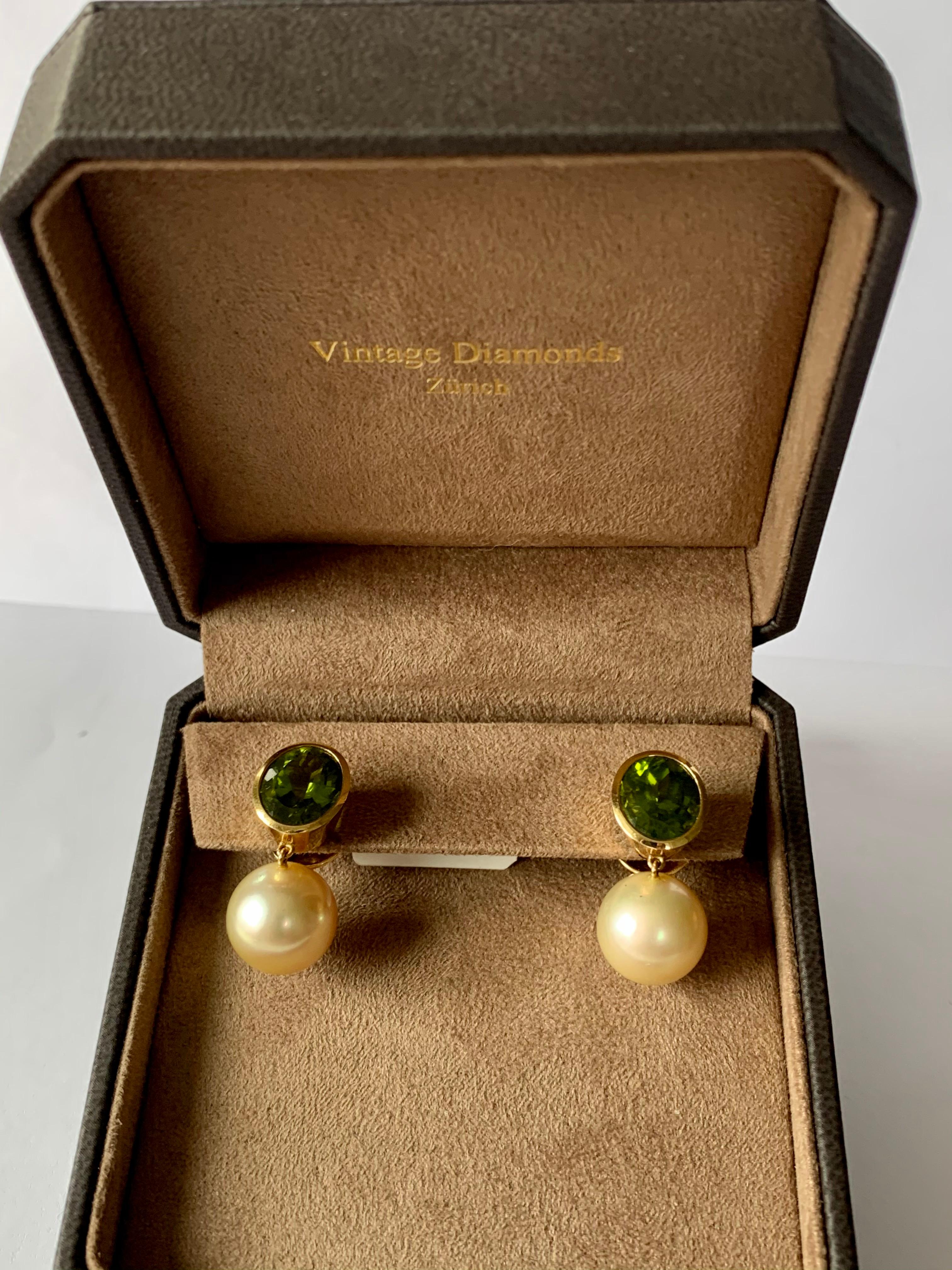 A pair of solid handcrafted 18 K yellow Gold earrings with 2 light golden South Sea pearls measuring 13.5 mm in Diameter. The top part features 2 bezel set fine Peridots totalling 11.47 ct.  A truly very nice and wearable pair of earrings with a