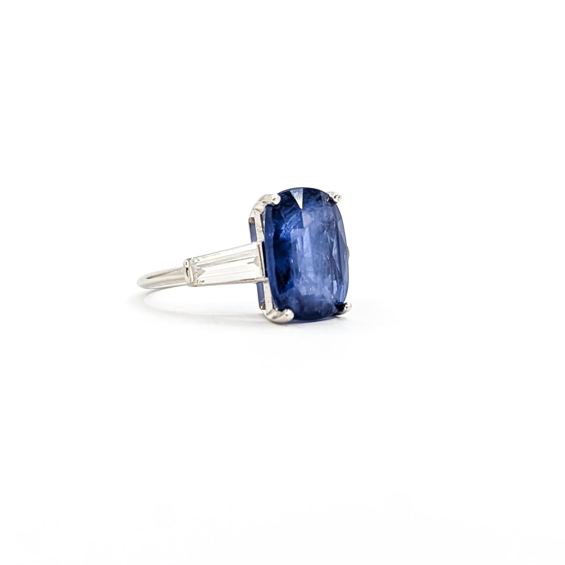 Cette bague classique est réalisée en platine et met en valeur un magnifique saphir bleu en serti clos pesant environ 6,01 carats. La pierre centrale est flanquée de deux diamants baguettes effilés totalisant 0,52 carats, presque incolores et d'une