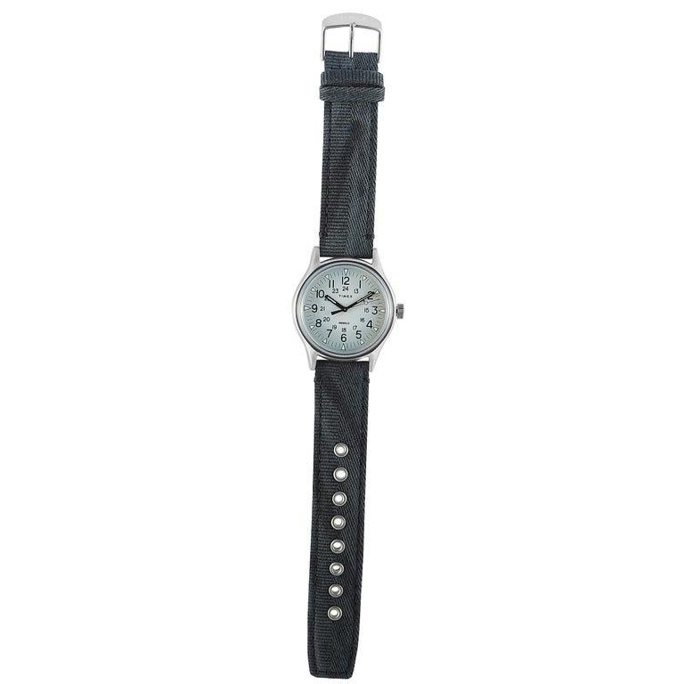 This is the Timex MK1 watch, reference number TW2R68300. It boasts a stainless steel case that offers water resistance of 30 meters. The case measures 40 mm in diameter and is presented on a gray fabric strap, secured on the wrist with a tang