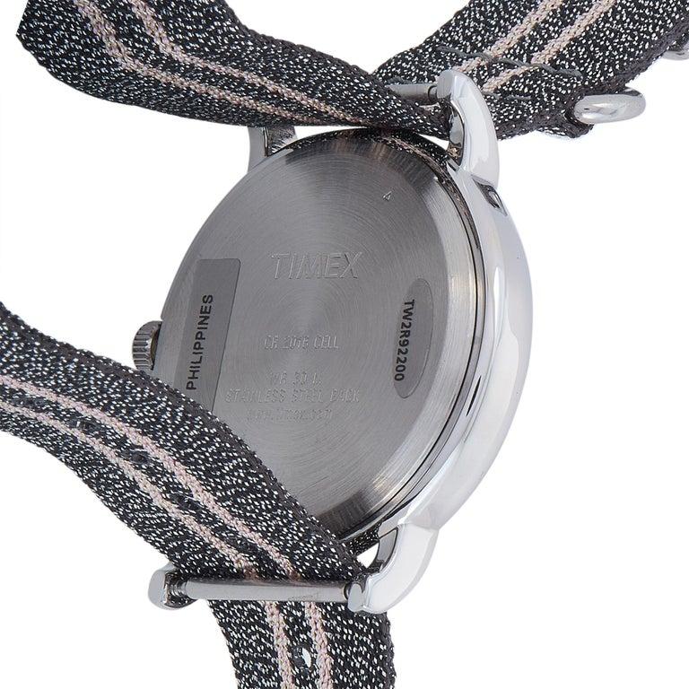 Dies ist die Timex Weekender 38 mm, Referenznummer TW2R92200. Die Uhr präsentiert sich mit einem 38-mm-Gehäuse aus silberfarbenem Messing und einem Boden aus Edelstahl. Das Gehäuse ist bis zu 30 Meter wasserdicht und wird an einem grauen,