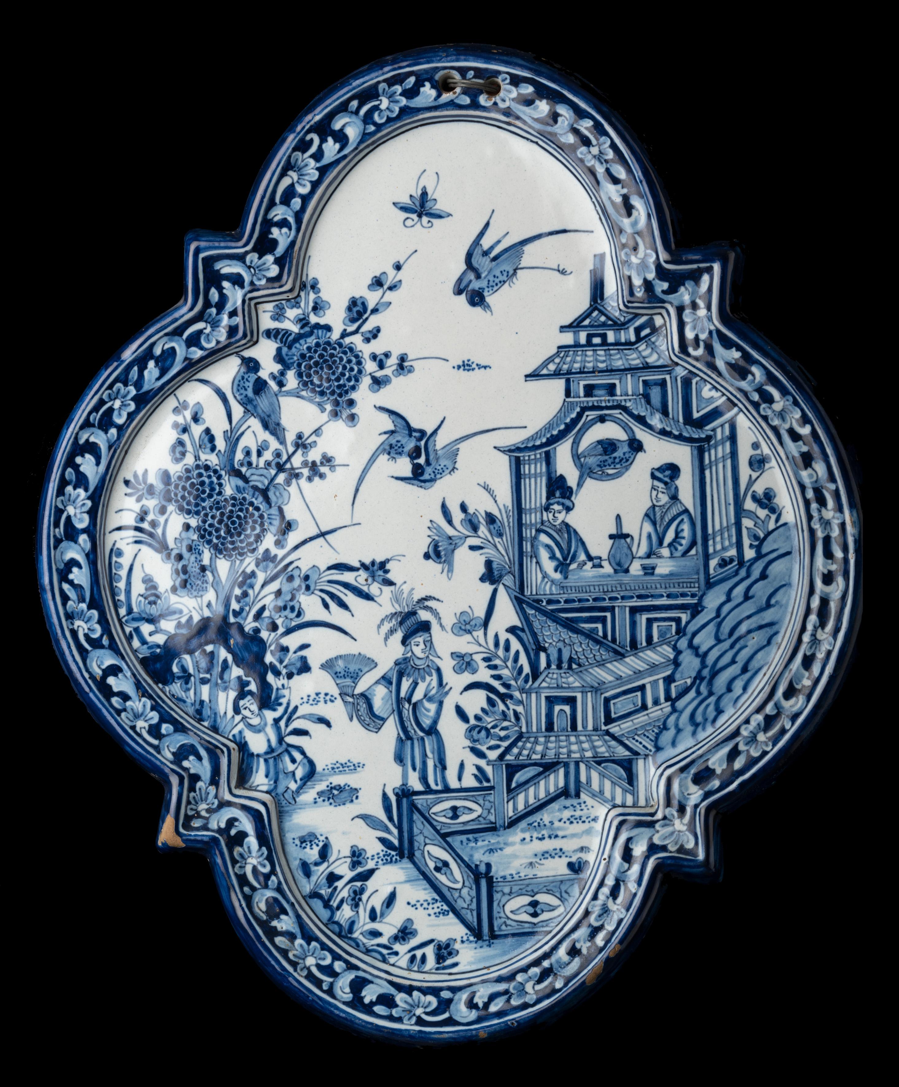 Tin-glazed plaque in the style of old Dutch Delftware, quatrefoil shape with raised rim, painted in blue with a chinoiserie decor of a pavilion in an oriental garden. Two figures are sitting in the pavilion, two more are in the garden. Birds and
