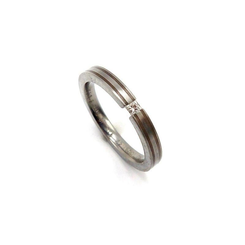 This lightweight yet strong titanium, silver, and diamond ring has a striking modernistic design. The band is made from titanium with two additional lines of inlaid silver that meet in the center around a tension fit square cut diamond — this adds
