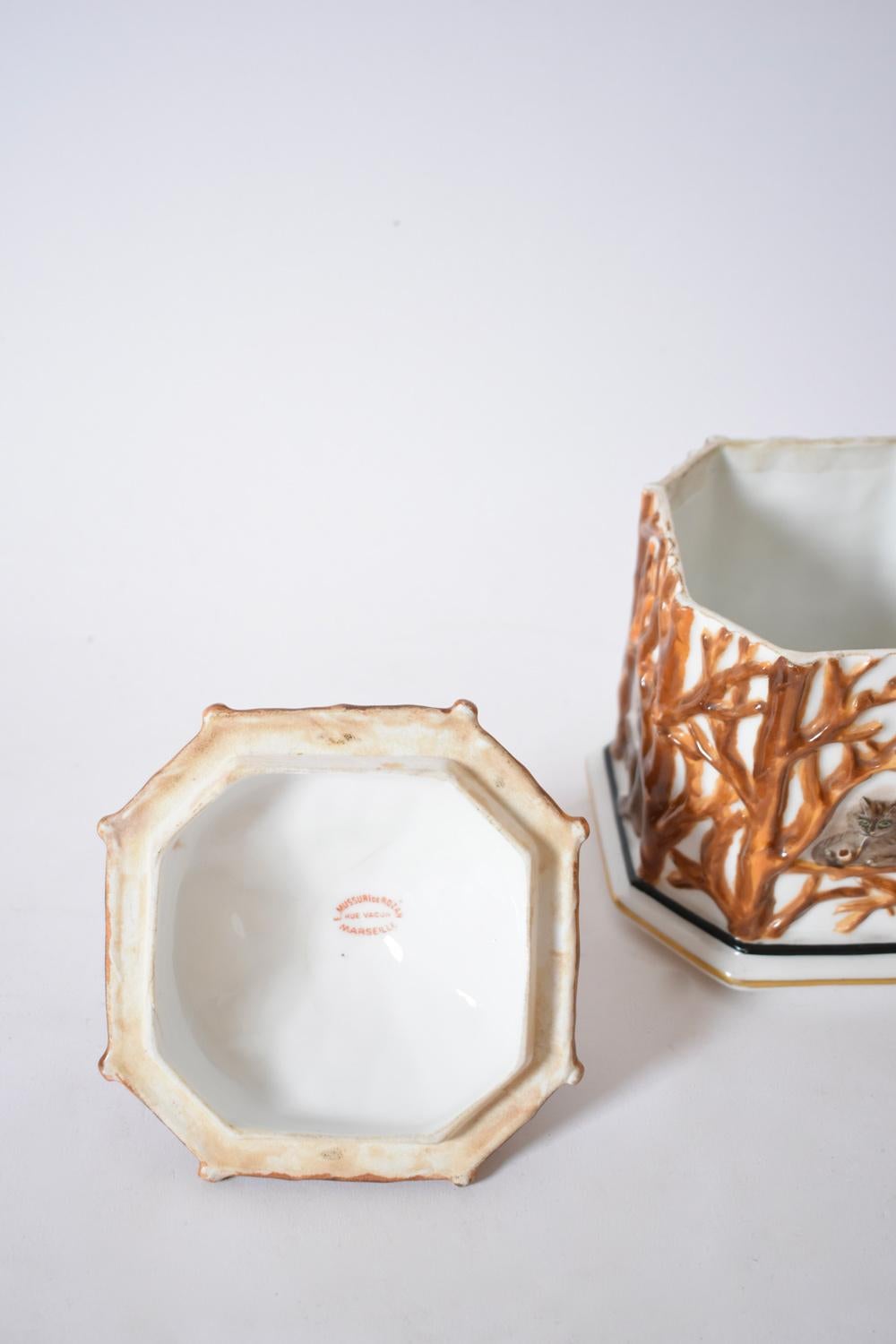 Pot couvert en porcelaine, à pans coupés, émaillé d'un décor en relief de feuillages bruns sur fond blanc, laissant apparaître sur chaque face un chien, un chat, un âne et un singe fumant. Le couvercle présente la même décoration et est surmonté