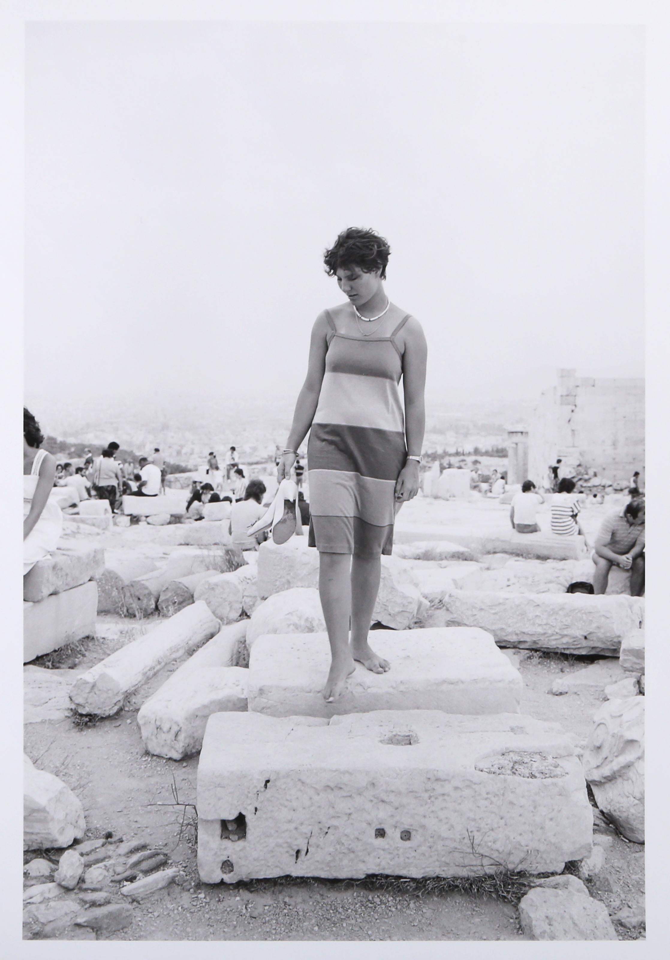 Woman with Shoes is a gelatin silver print from On the Acropolis, a portfolio of 25 photographs by Tod Papageorge made on the Athenian Acropolis in the summers of 1983 and 1984. The image depicts a young woman in a striped sleeveless dress standing