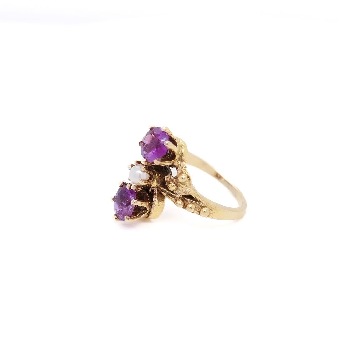 A fine toi et moi gold, amethyst, and pearl cocktail ring.

Size 2.

In 14 karat yellow gold.

The gold setting with a flower to either side of the stones and with decorative rivets/beading.

Set two round cut amethysts with a pearl in the center of