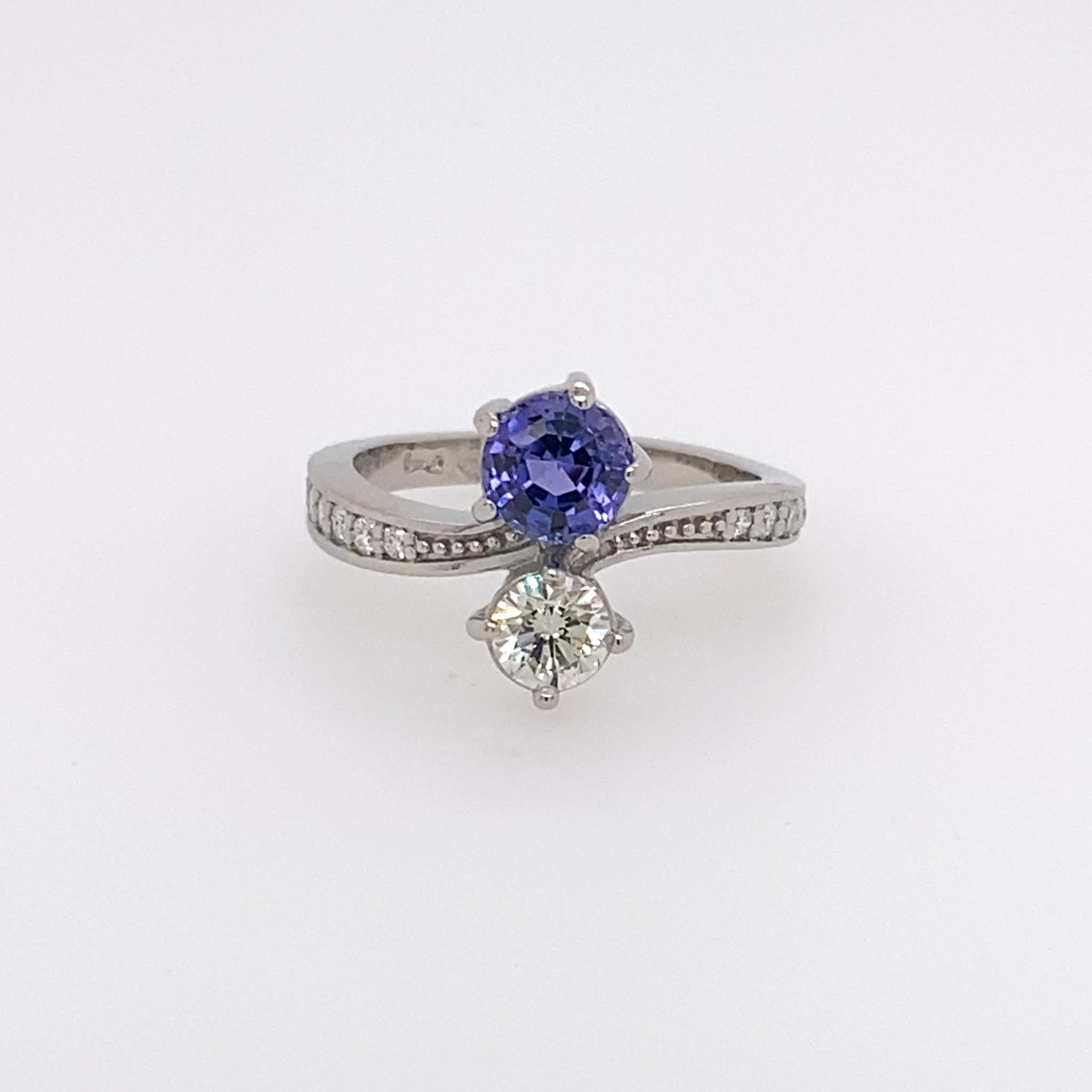 Classic design containing a round modified step cut Tanzanite secured in a five claw setting and round brilliant cut diamond  in four claw setting. The two stone are opposing one another. The curved shoulders each house round brilliant cut diamonds