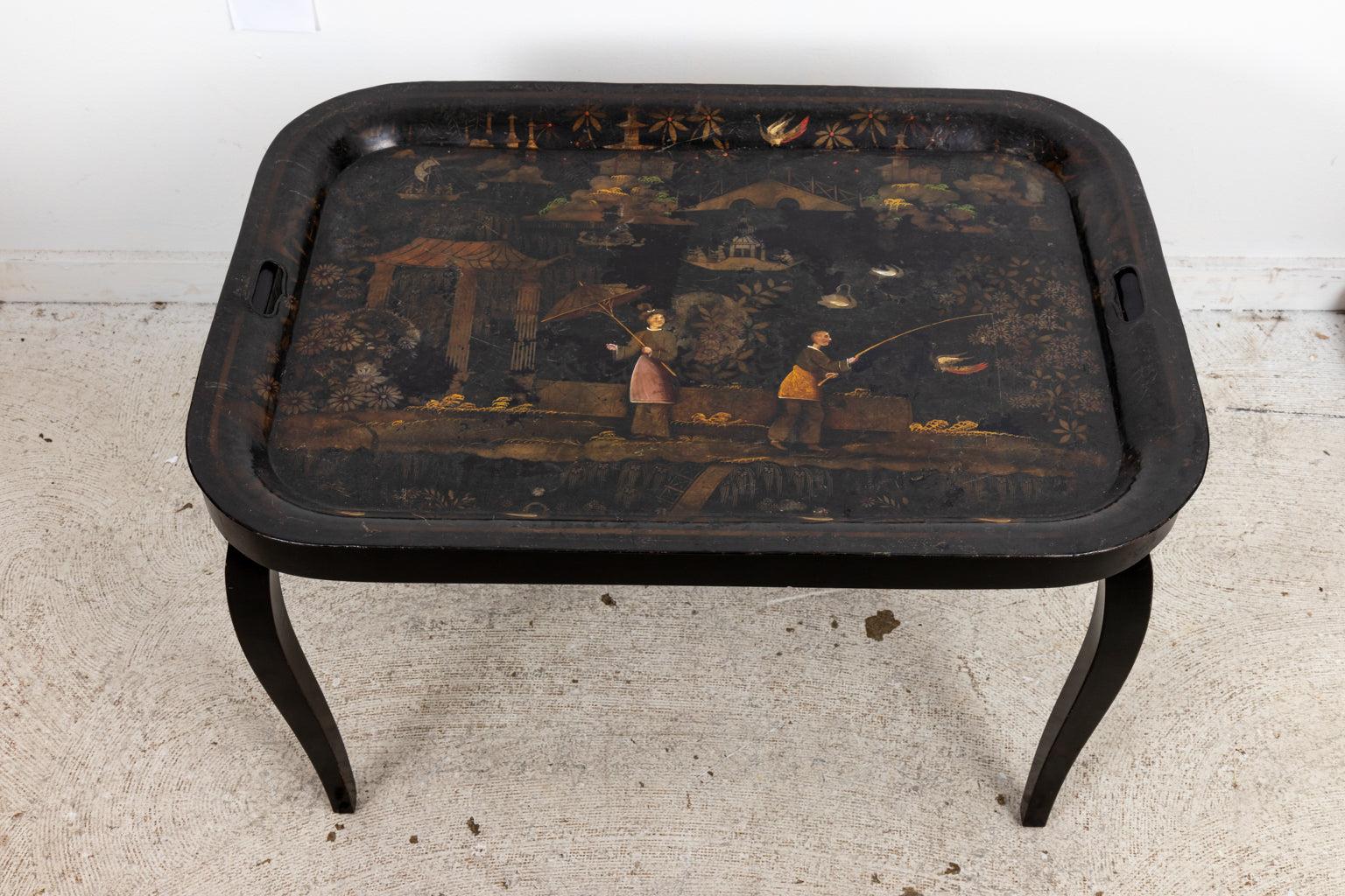 Circa 1920s black tole Chinoiserie style tray table with a custom base and a painted village scene on the tabletop. Origin unknown. Please note of wear consistent with age including minor chips, paint loss, patina, and finish loss.