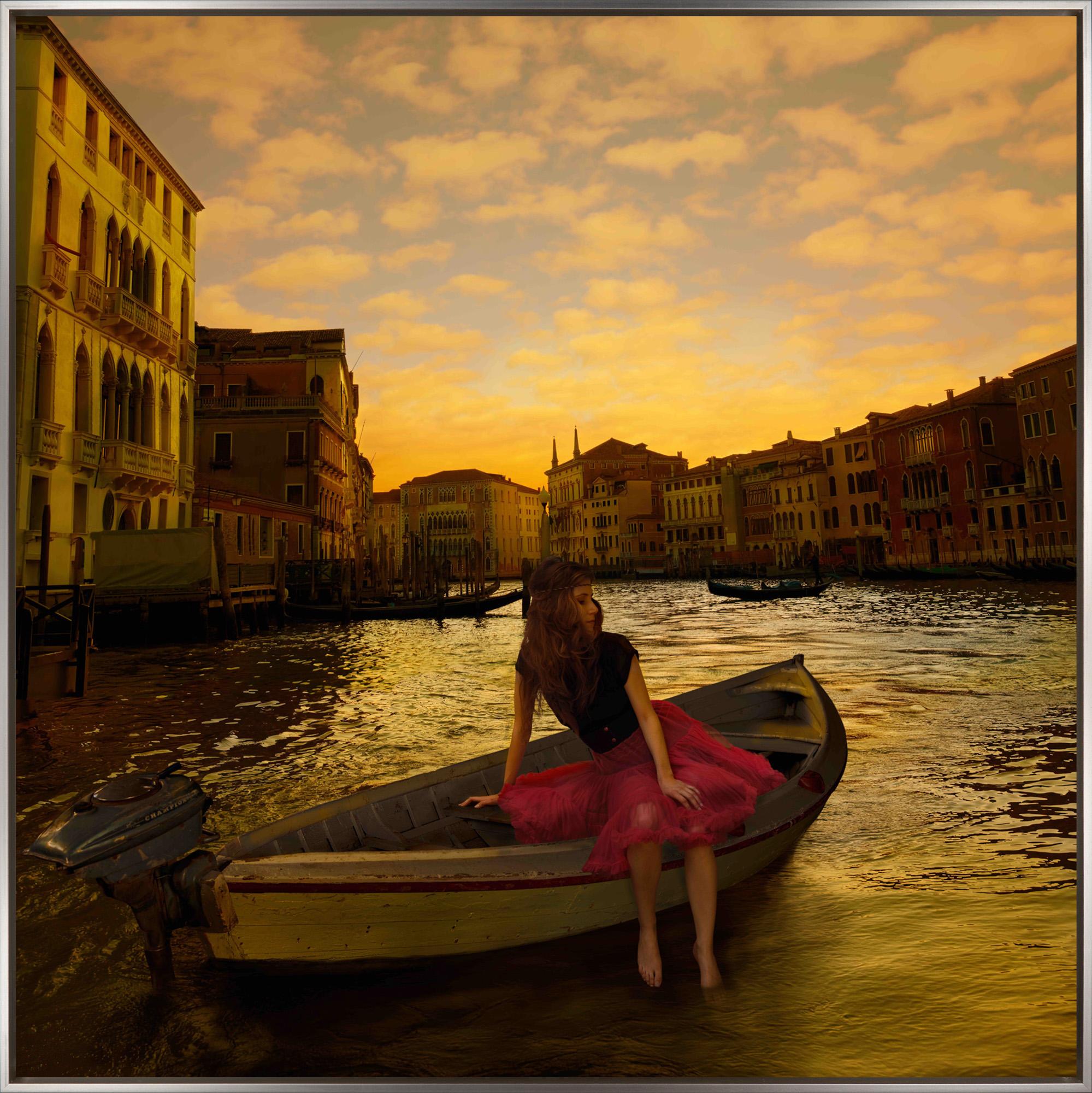 "Morning on the Grand Canal" is a framed photograph on aluminum by Tom Chambers, depicting a young woman in a small boat adrift in Venice. The crisp finish of the printed photograph is balanced by the warm color palette, evoking a dreamlike quality