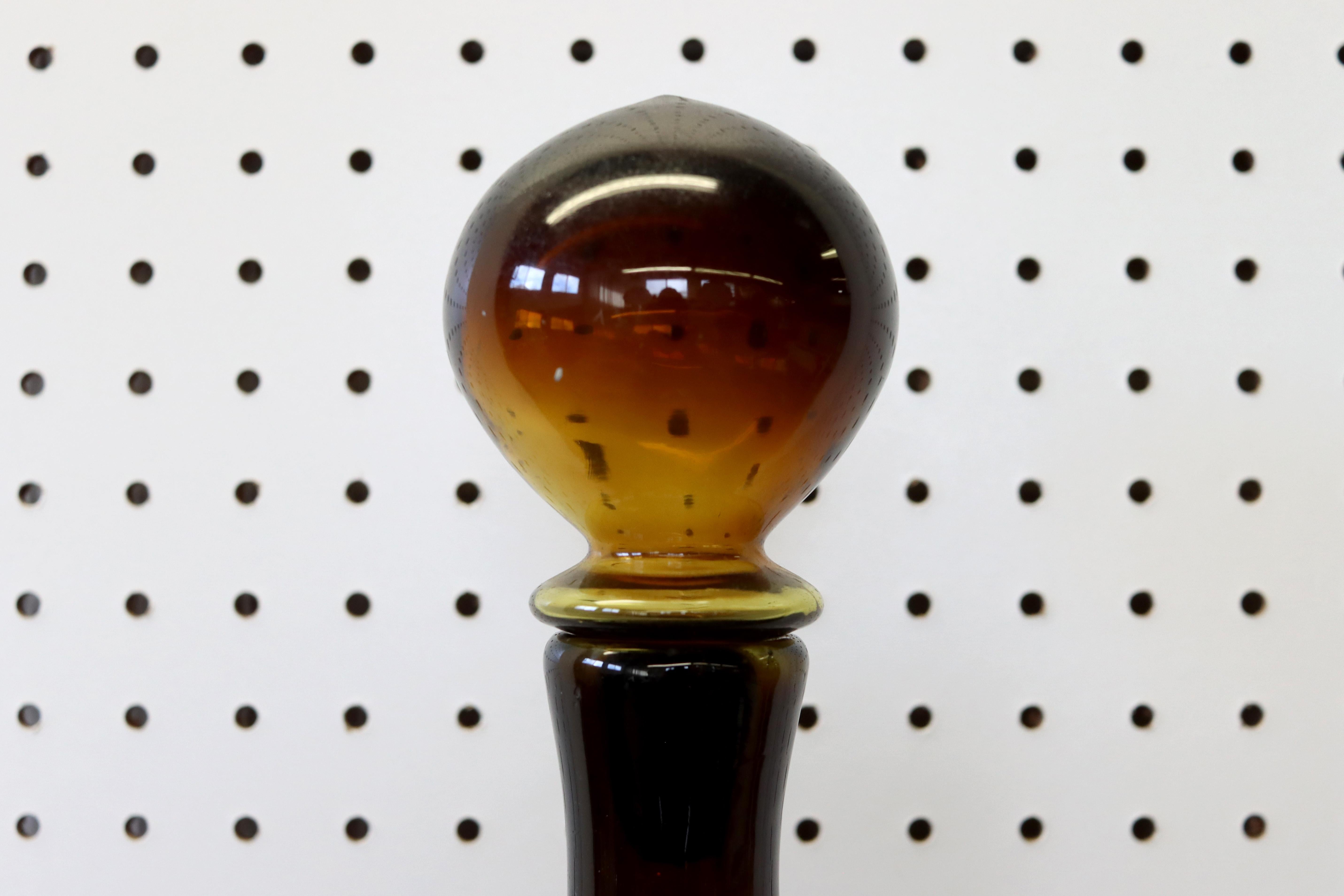 A burnt-honey glass decanter by Tom Connally for Greenwhich Flint-Craft. Made circa the late 1960s. Includes original stopper. The decanter is in excellent condition with no chips, cracks, or clouding. Measures 25 1/4” tall x 3 7/8” diameter.