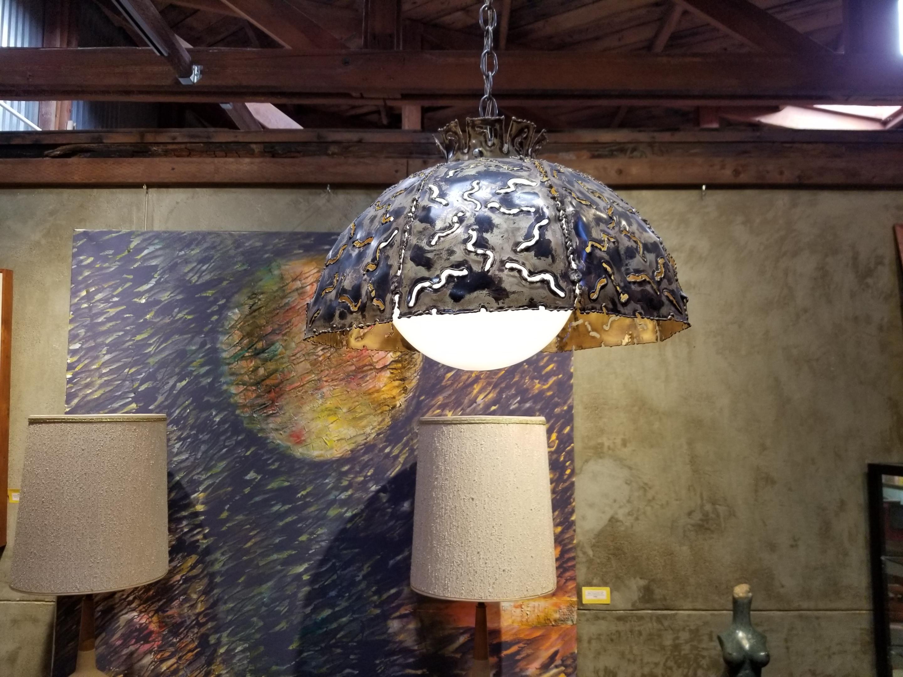 A Tom Greene torch cut brass Brutalist lighting fixture by The Feldman Company. Features a milk glass shade to diffuse glare. Hangs as a swag with a 15 foot chain or may be installed as a chandelier by omitting most of the chain and using an