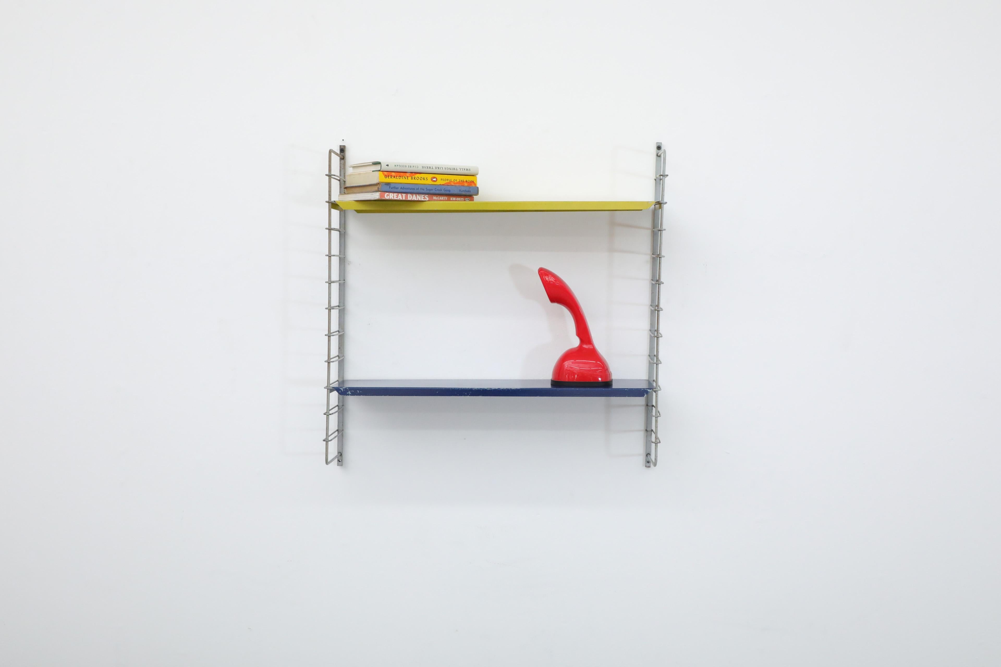 Single section Tomado style wall mounted shelving unit with yellow and blue metal shelves on thin light gray wire risers. In original condition with visible wear consistent with its age and use. Similar shelving units also available and listed