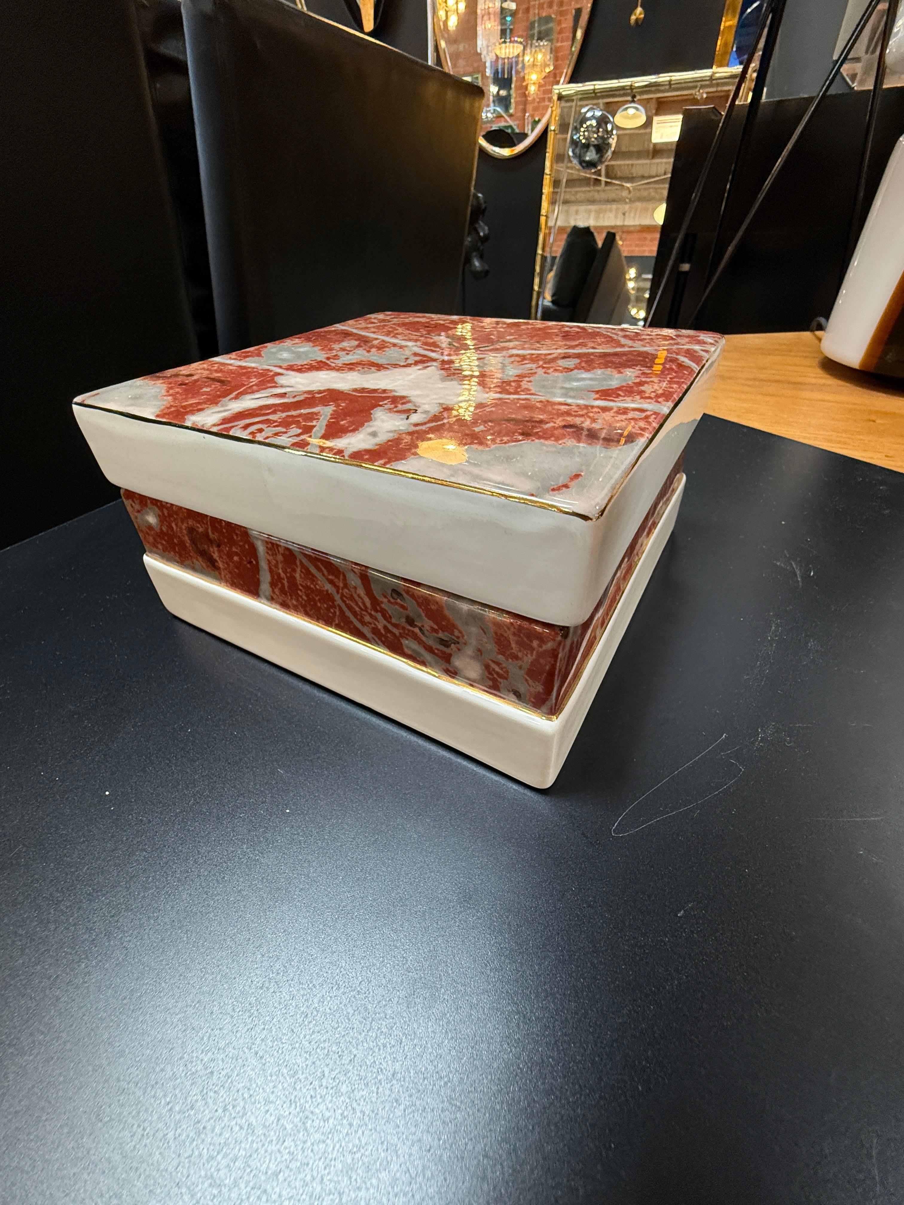 Esta elegante caja con tapa de Tomaso Barbi muestra su característica mezcla de formas escultóricas y refinados detalles decorativos. La caja cuadrada está fabricada en cerámica, acabada con un llamativo esmalte marmoleado rojo y gris, acentuado por
