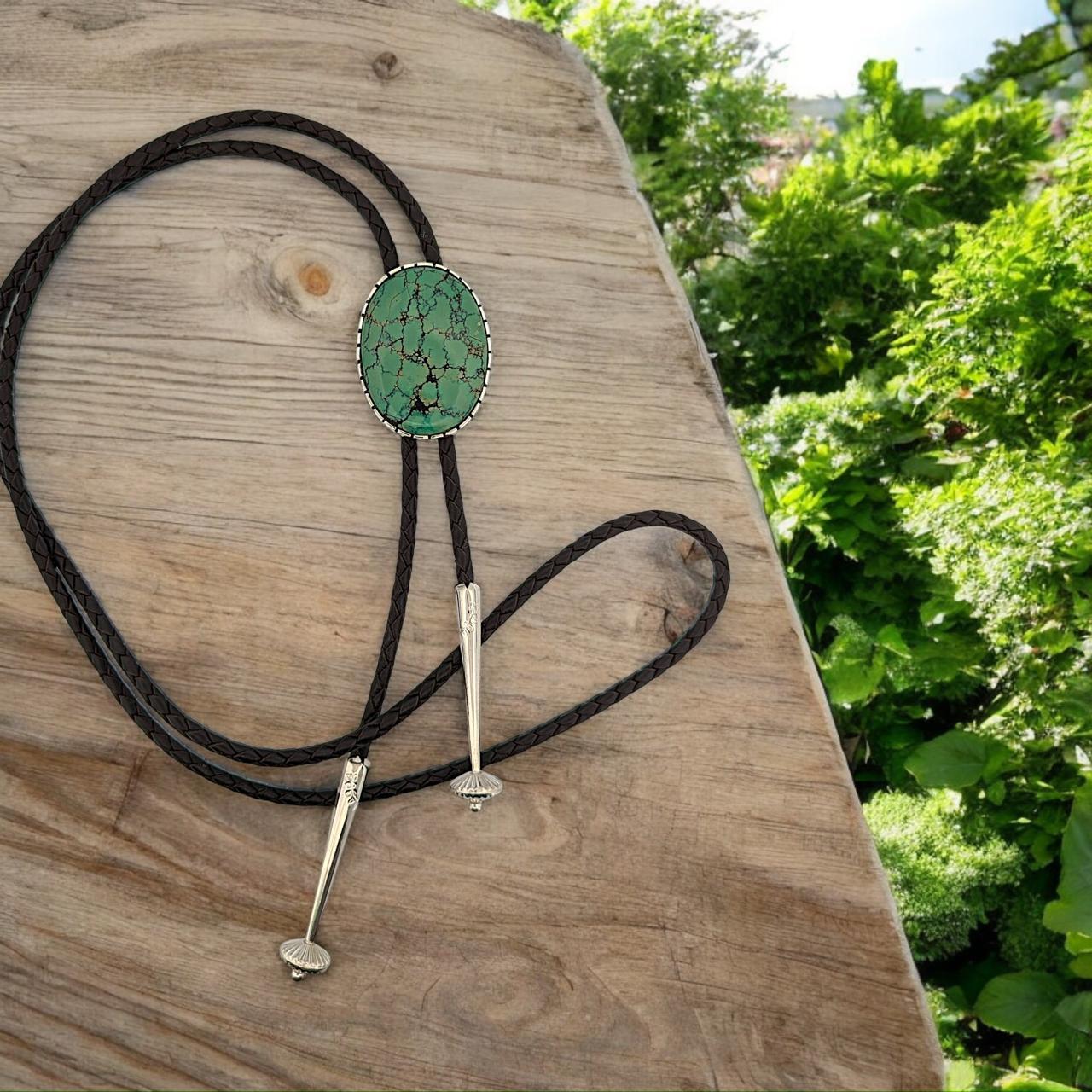 Introducing Tombstone Bolo, a handcrafted bolo tie by Rob Sherman that features a stunning green Kingman turquoise cabochon with a rich chocolaty brown matrix. Set in a classic high polish bezel, this exquisite bolo tie is designed with sleek,