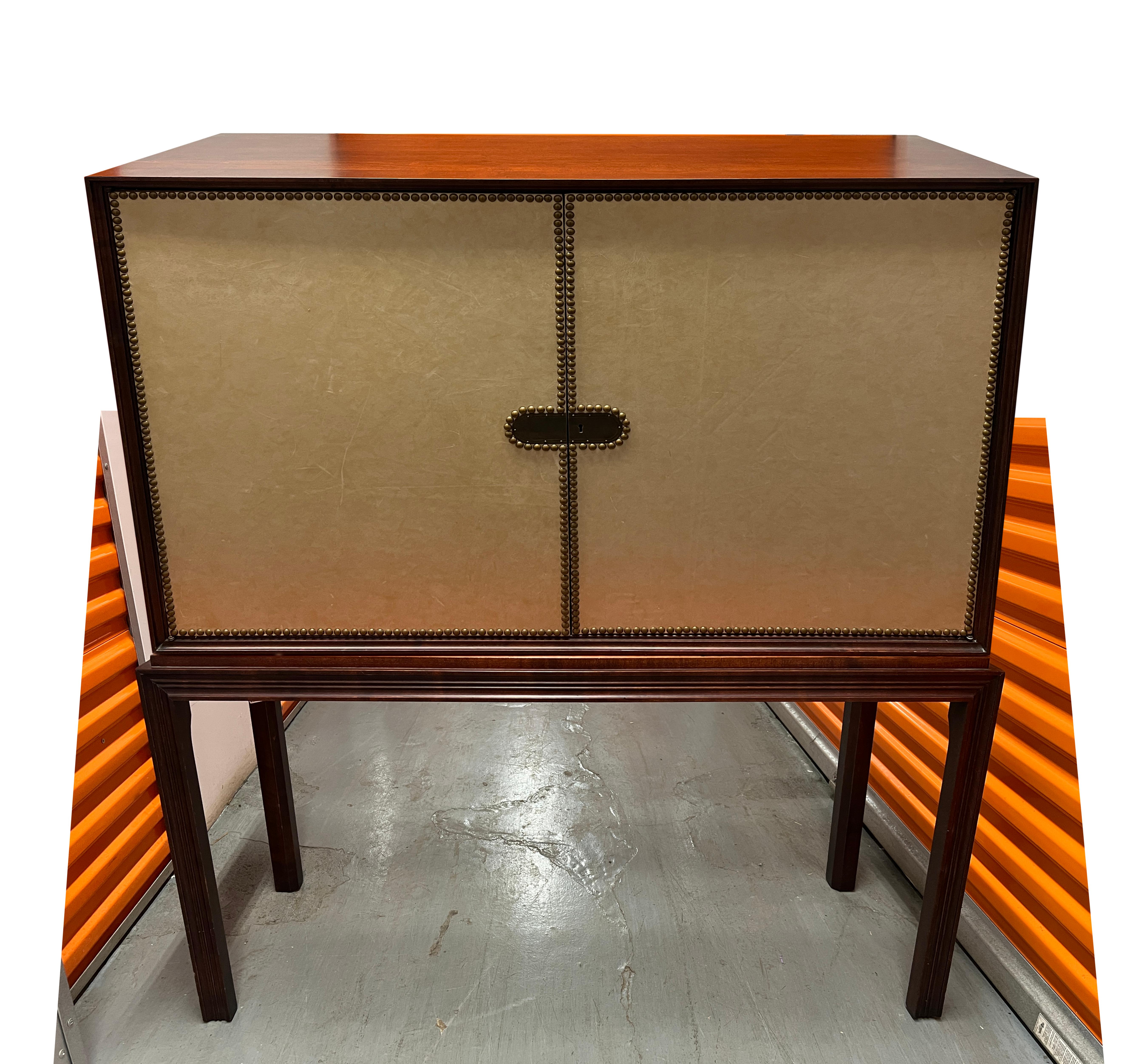Tommi Parzinger for Charak Modern chest of drawers on stand in mahogany with leather-front doors and brass nailheads. Cabinet has two doors with lock and eight interior drawers. Manufacturer's label ‘Charak Modern’ inside of drawer. Key for lock