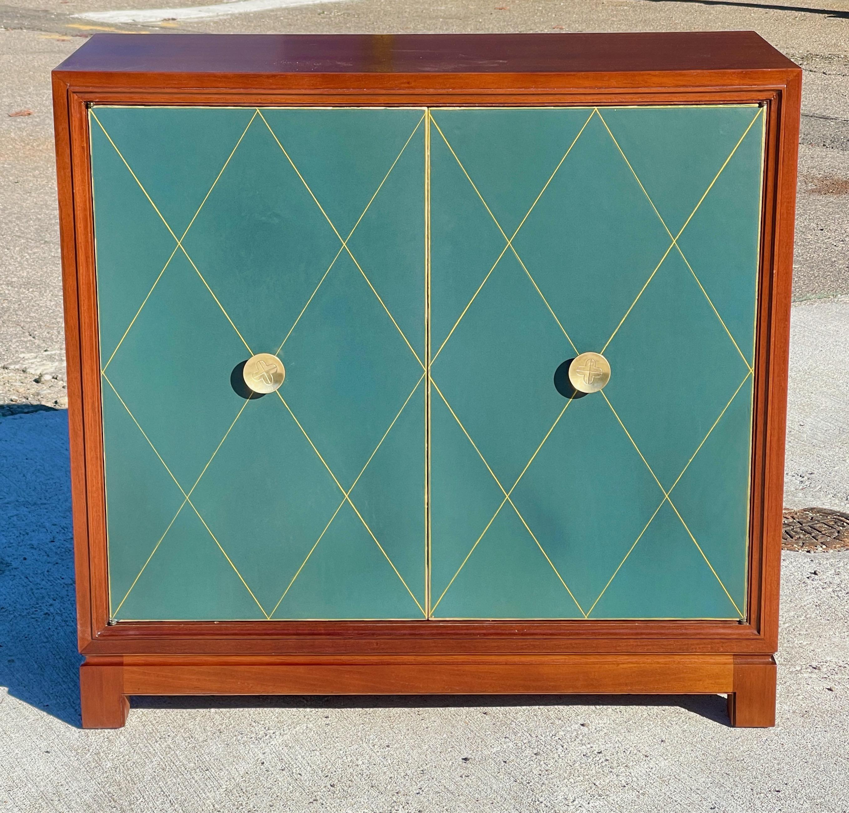 Classic mahogany cabinet with tooled and gilt diamond pattern green leather door panels and distinctive polished brass door pulls. 
Features a single adjustable interior shelf. 
Designed by Tommi Parzinger and produced by Charak Modern in 1953.