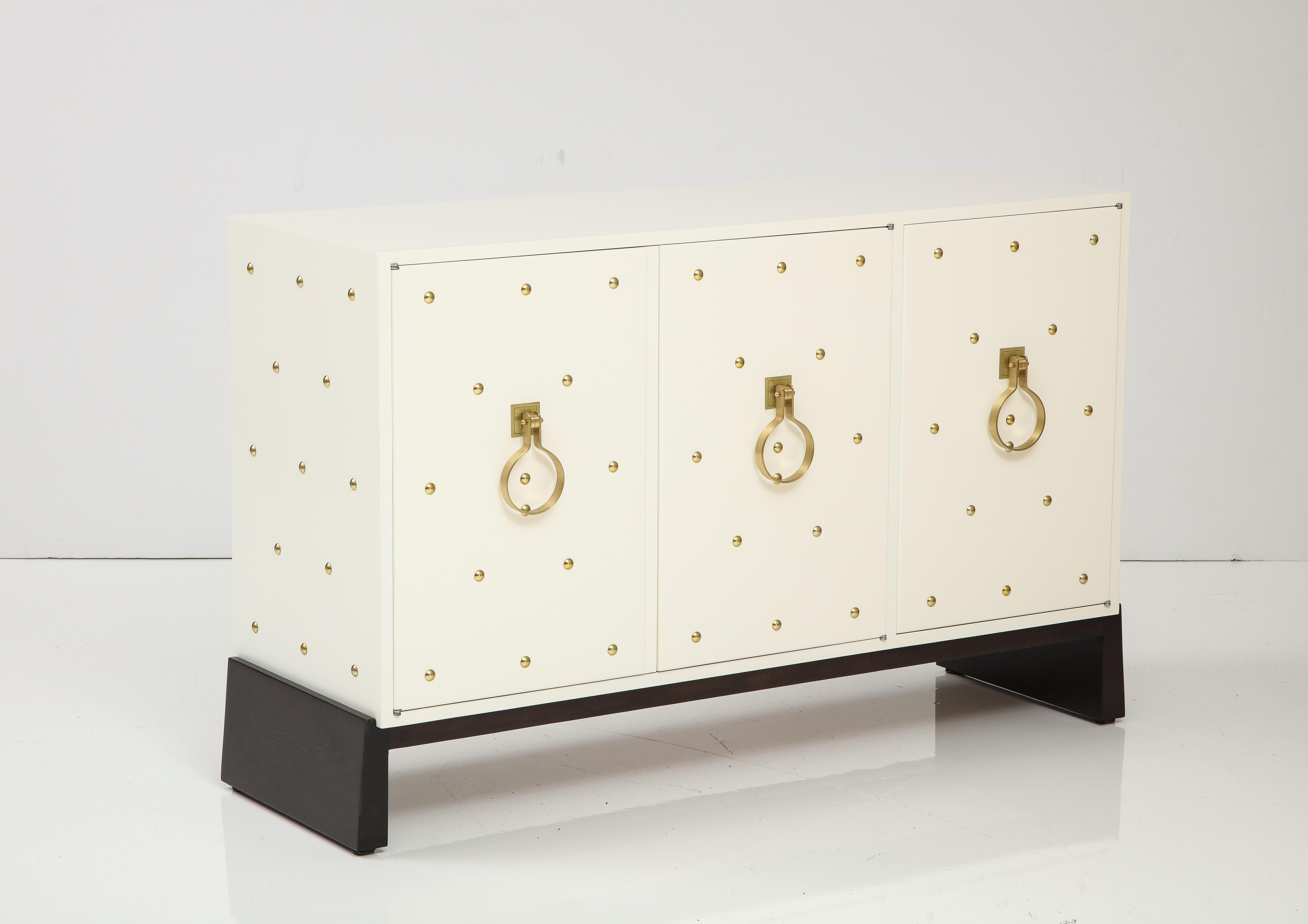 Tommi Parzinger for Parzinger Originals three door studded cabinet. 
The cabinet has been Newly restored in a soft white Matte finish and brass studs and sits on an espresso colored plinth base.
The interior has a single interior drawer with the