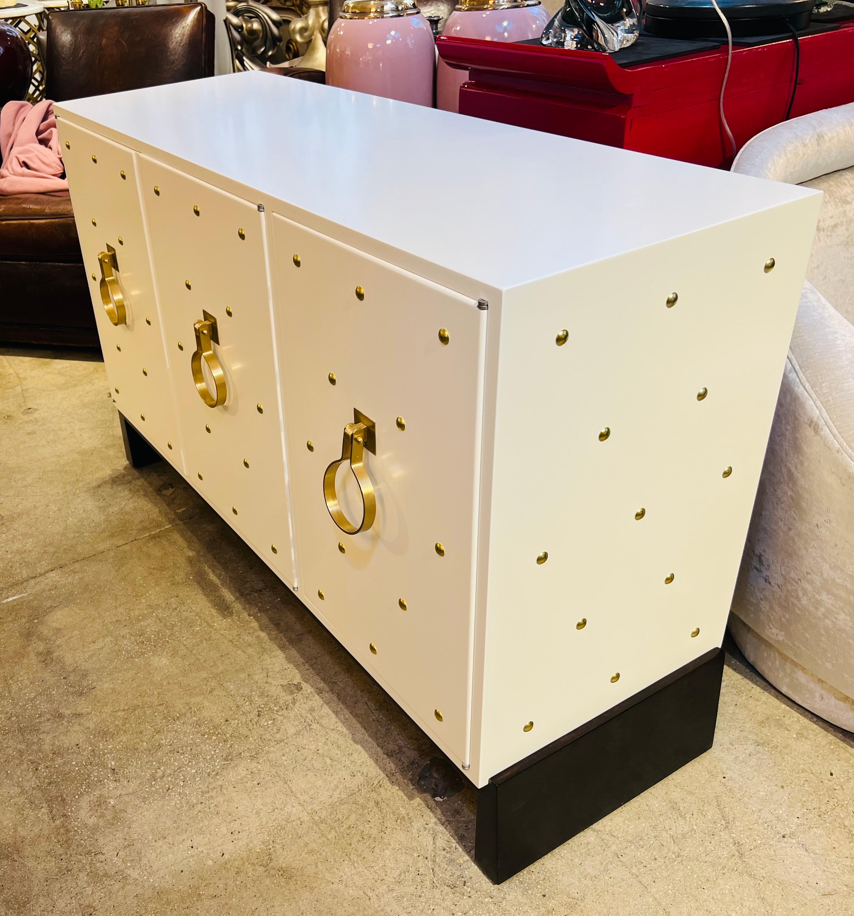 A three door matte white cabinet on a espresso plinth base. The sideboard cabinet has beautiful satin brass pulls and studs to the face and sides. Drawers and shelves are held inside. Excellent beautiful condition. Signed with the makers mark.