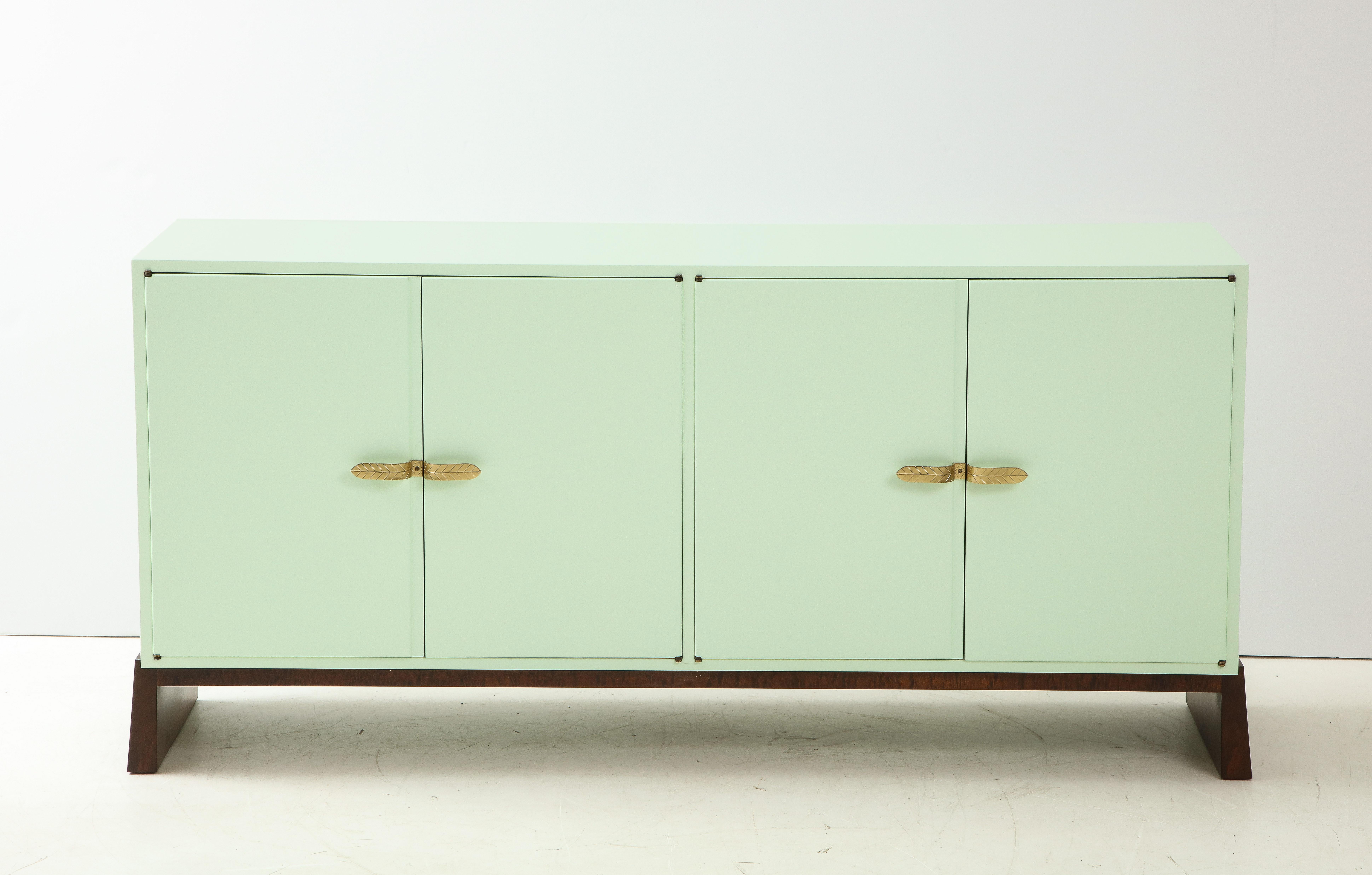 American midcentury sideboard featuring a seductive matte finished Pistachio color lacquer with stylized brass leaf handles, interior finished in a white matte lacquer with shelves. Cabinet rests on splayed walnut base. Stamped Parzinger Originals.