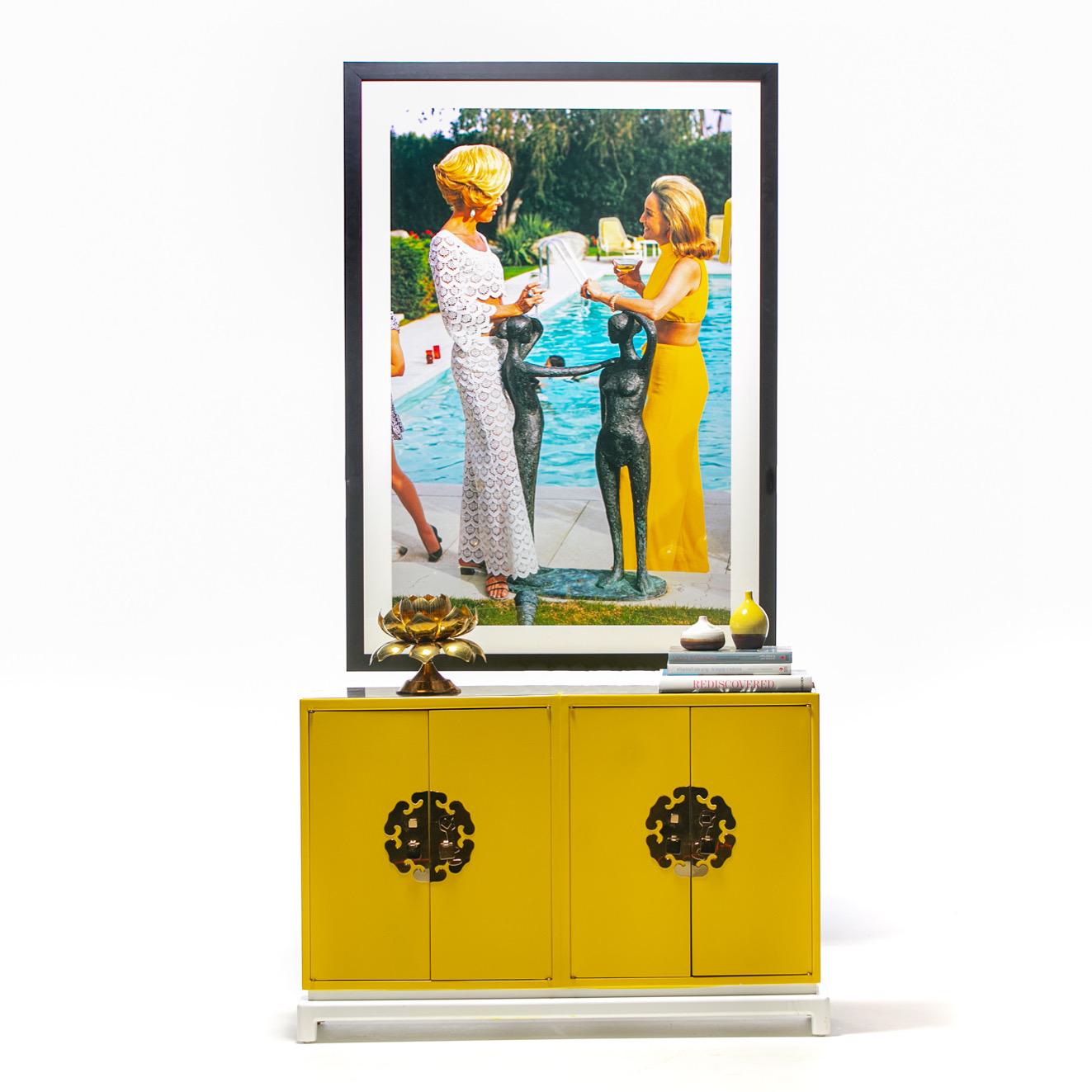 1960s Hollywood Regency cabinet or console professionally refinished in yellow and white lacquer with original Chinoiserie medallion hardware and mirror top in the style of Tommi Parzinger resting atop a single pedestal wood frame. Brass Medallions