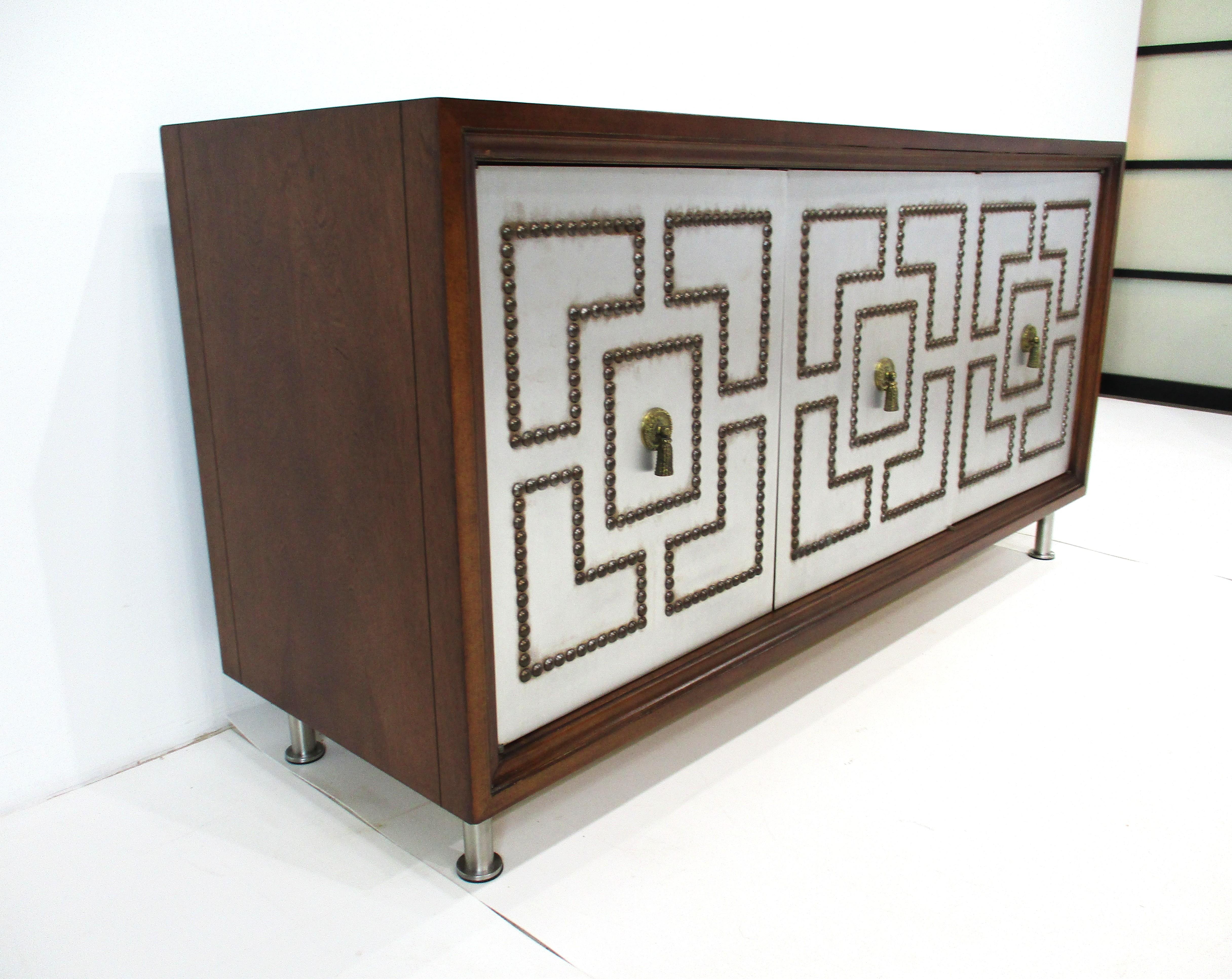A unique Mid Century dark walnut three door credenza with applied metal fronts having brass studs and cast brass tassel shaped pulls. Inside is one fixed shelve running the length of the cabinet with brassy molted painted patterns to the inner