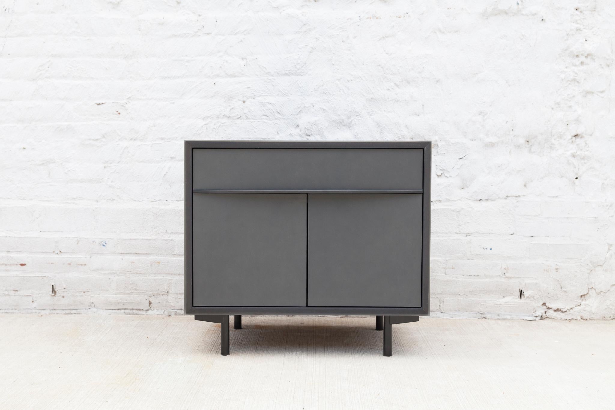 The sleek Tompkins end table has a blackened aluminum encased in epoxy resin exterior. The doors and drawer face are wrapped in leather with blackened steel pulls. The walnut dovetail drawer is lined with black leather and the interior cabinet has