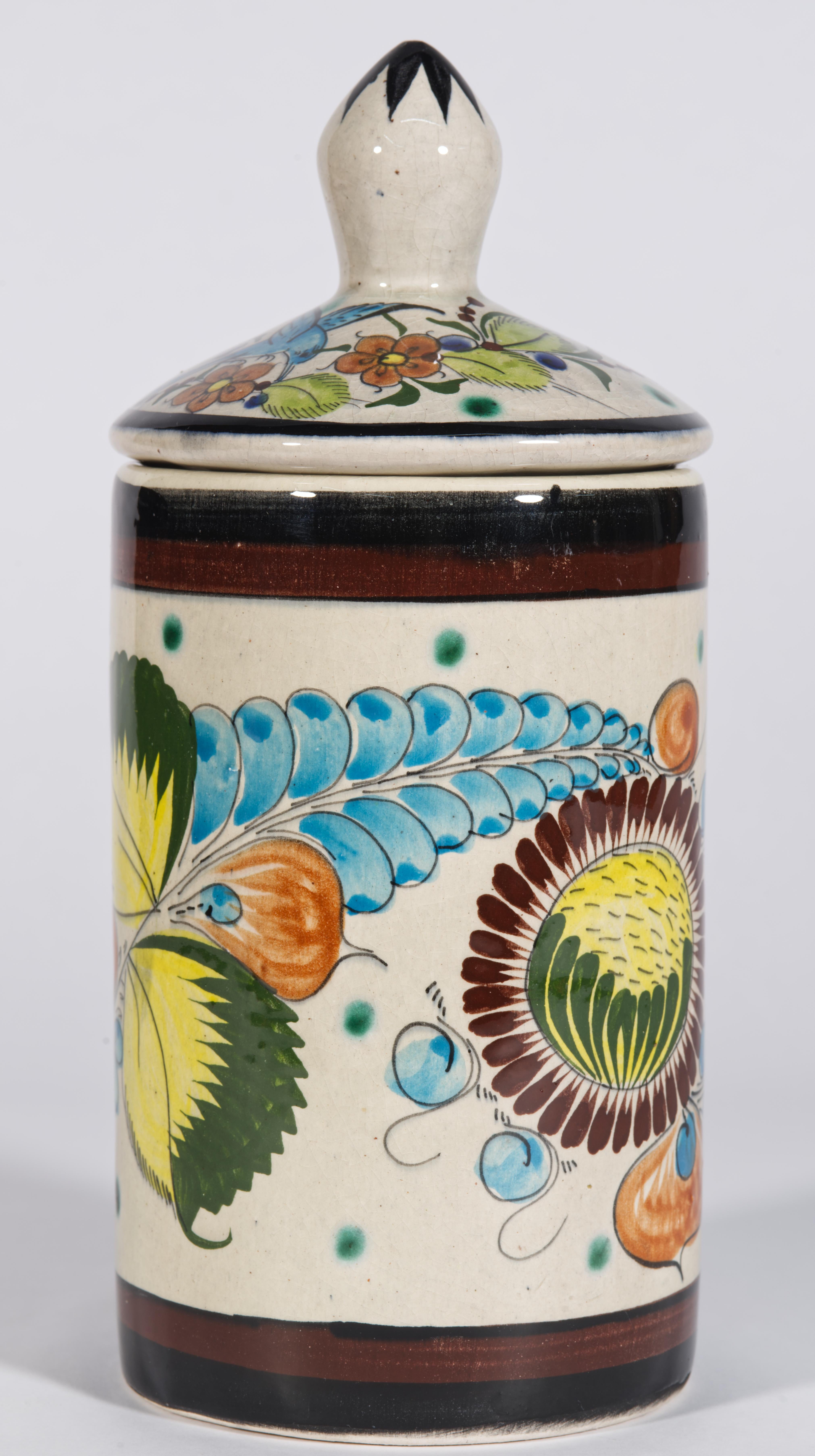 El tarro de cerámica en forma de cilindro presenta una decoración de estilo Folk Art con motivos florales y guirnaldas muy detallados, que se repiten en la parte delantera y trasera del tarro. La tapa está decorada con un pájaro azul que vuela sobre