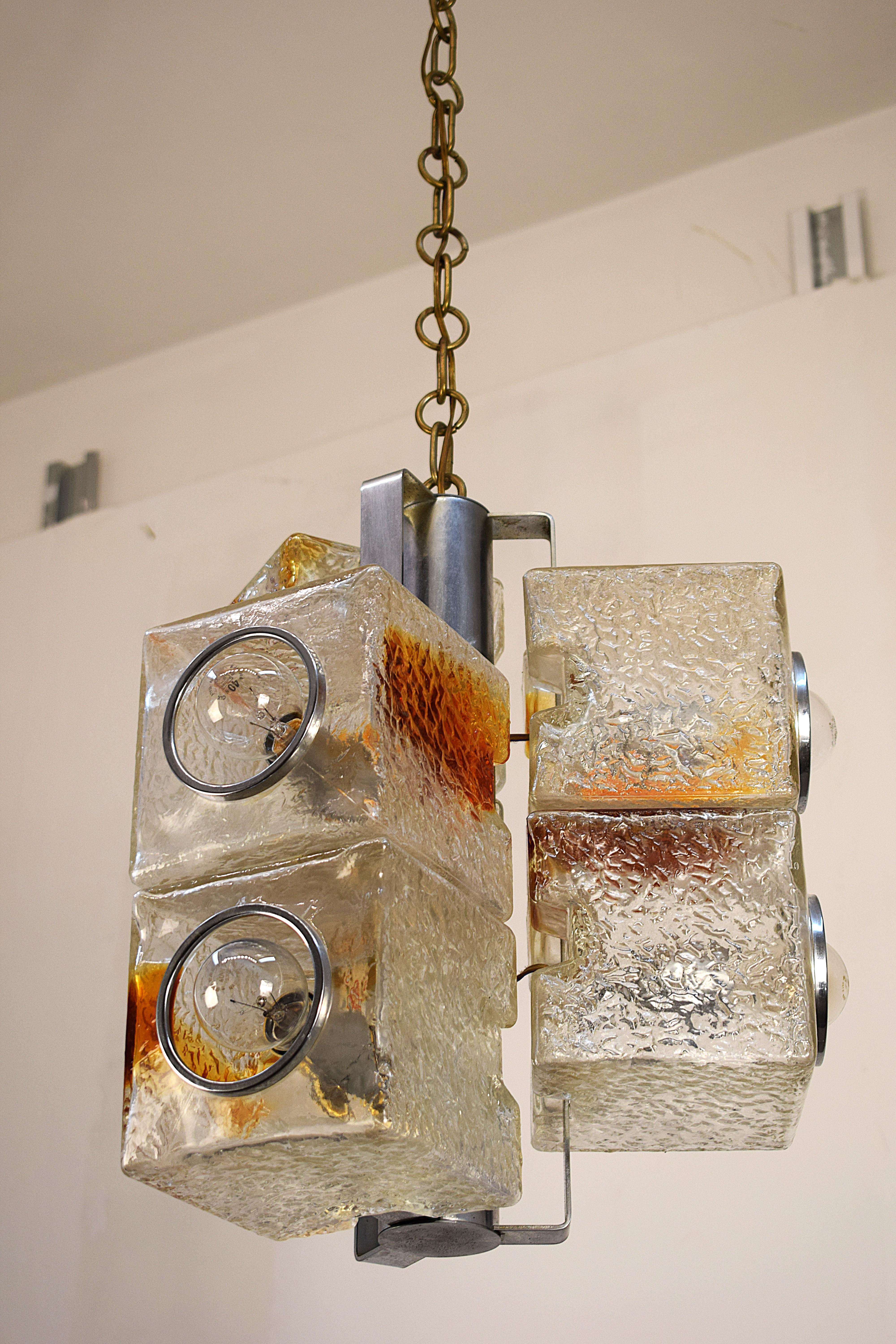 Affascinante pezzo di design italiano realizzato in acciaio e vetro di Murano, questo lampadario è un esempio autentico dello stile degli anni 1970. Lavorato con vetro spesso e decorato con tonalità ambrate, dona un tocco elegante e artistico a