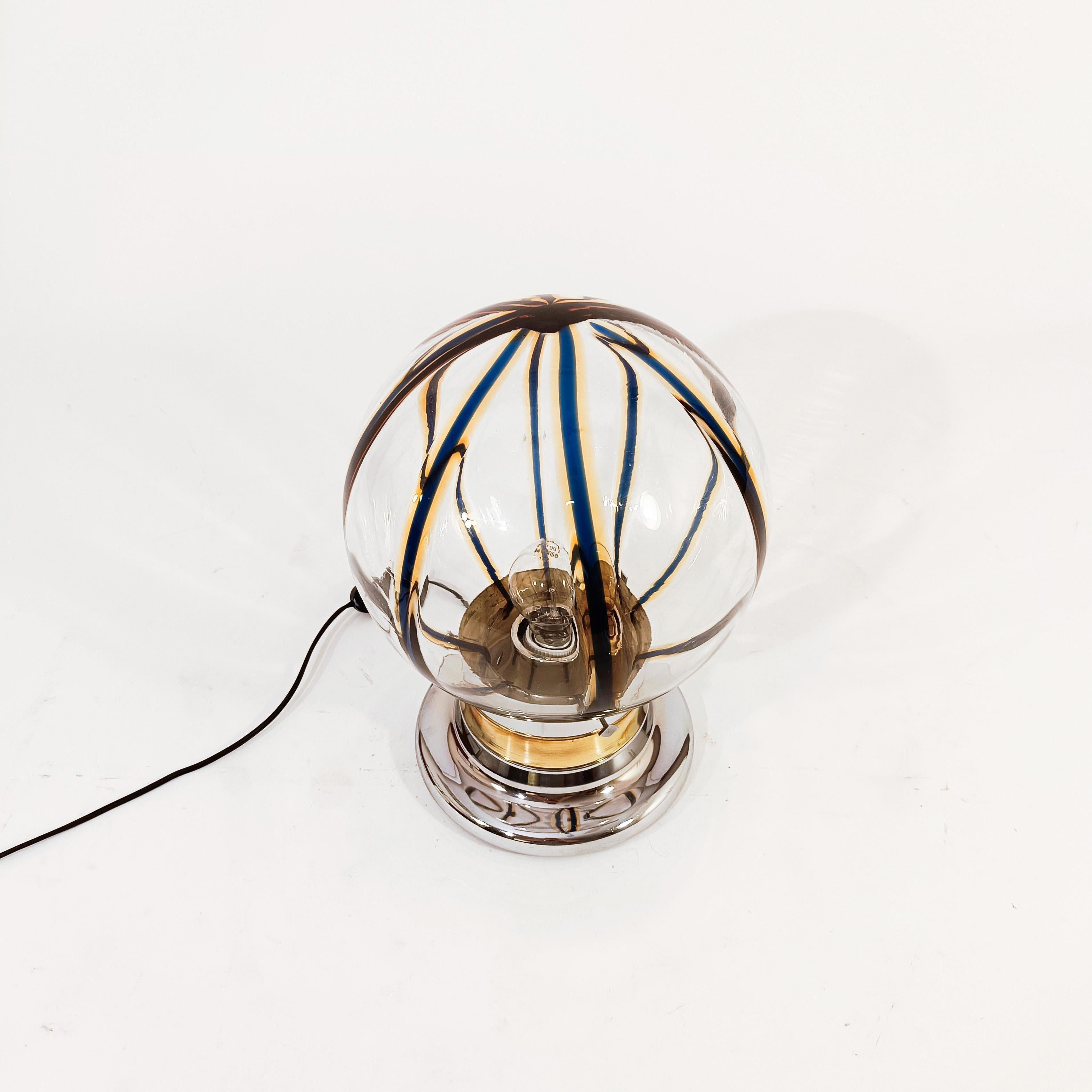 Italian brutalist and Space Age style table lamp by Toni Zuccheri, 1970s. Golden and silver chromed steel metal base and a spherical upper diffuser made of transparent Murano glass combined with navy blue and amber stripes, which provide a light