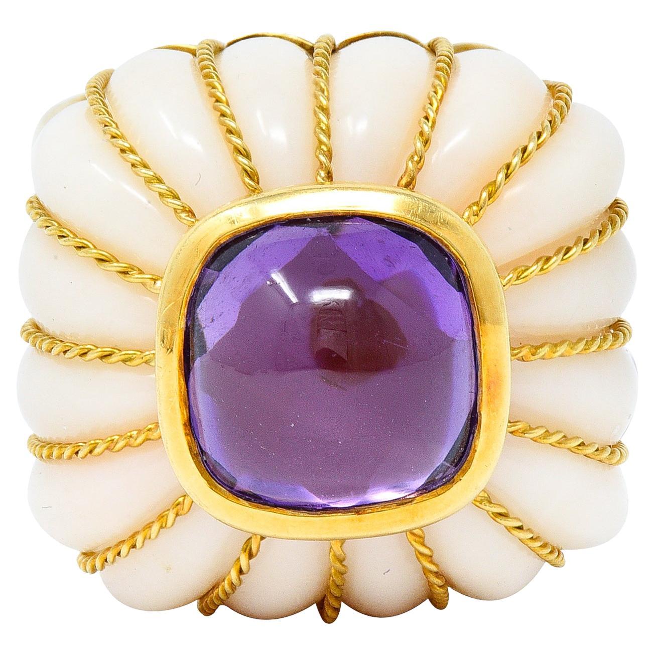 Centering a cushion shaped amethyst cabochon measuring 10.5 x 10.5 mm - transparent medium purple. Bezel set atop a cushion shaped coral - carved with fluted ridges fully around. Translucent cream in body color - ridges feature twisted rope motif