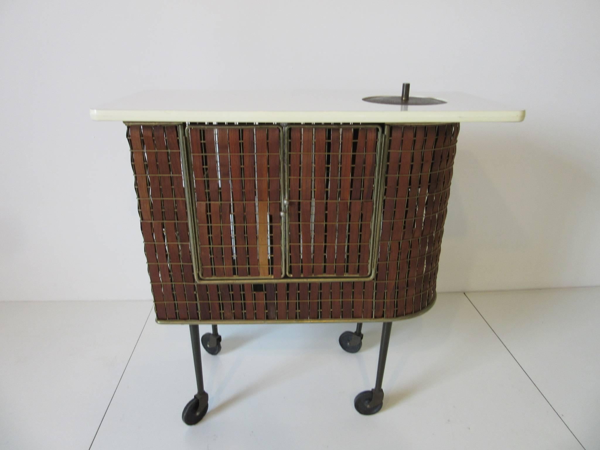 A early Tony Paul rolling bar cart called a Barmobile with white Formica top, built - in three quart ice bucket with lid and brass toned frame and teak woven strips. The backside double doors having a magnetic latch with storage inside , racks for