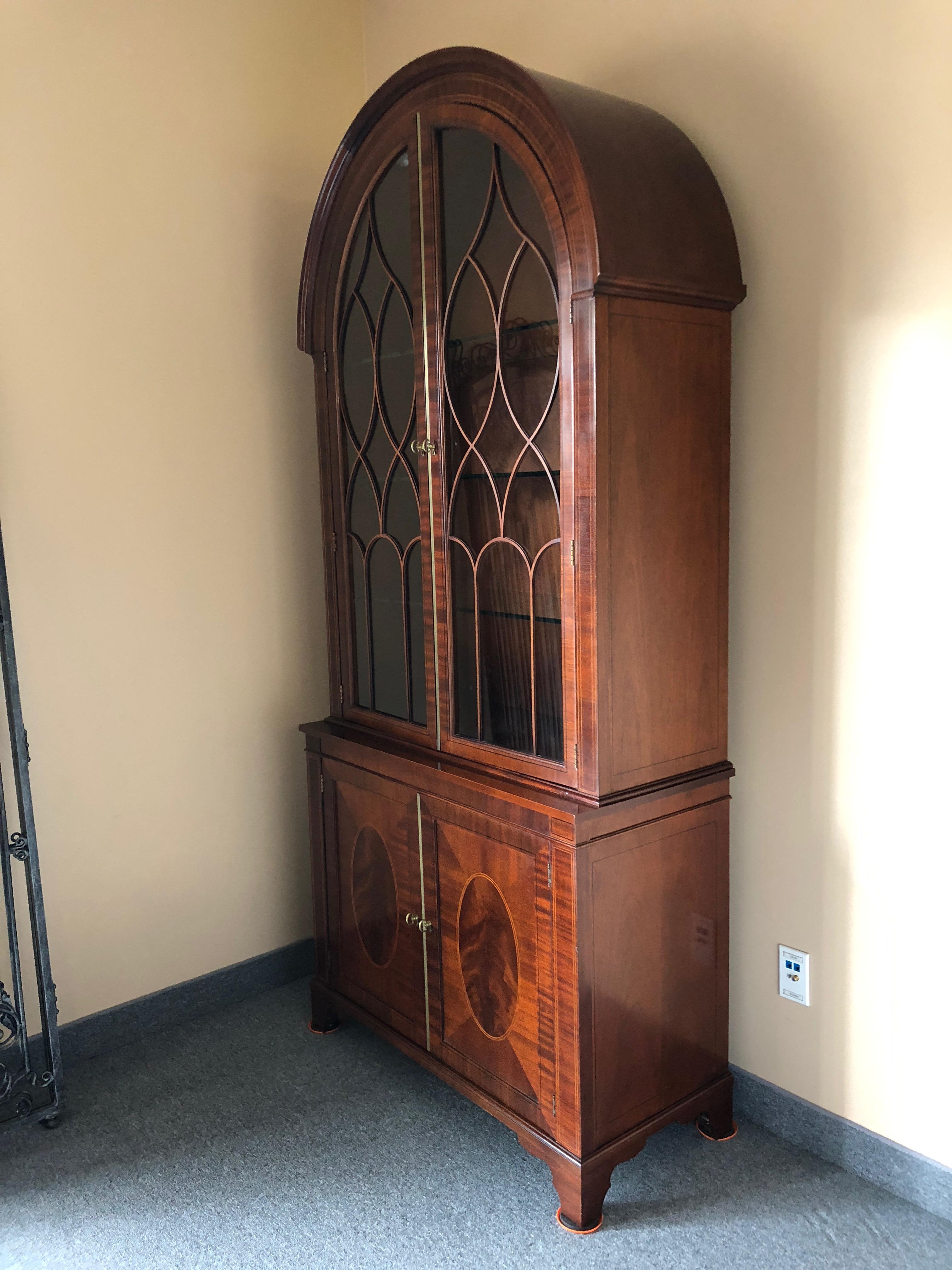 Beautifully made very elegant Baker breakfront or china display cabinet having a lovely rounded arch shape with glass front decorated with pretty criss crosses of wood, interior adjustable glass shelves, and flame mahogany bottom having ebony and
