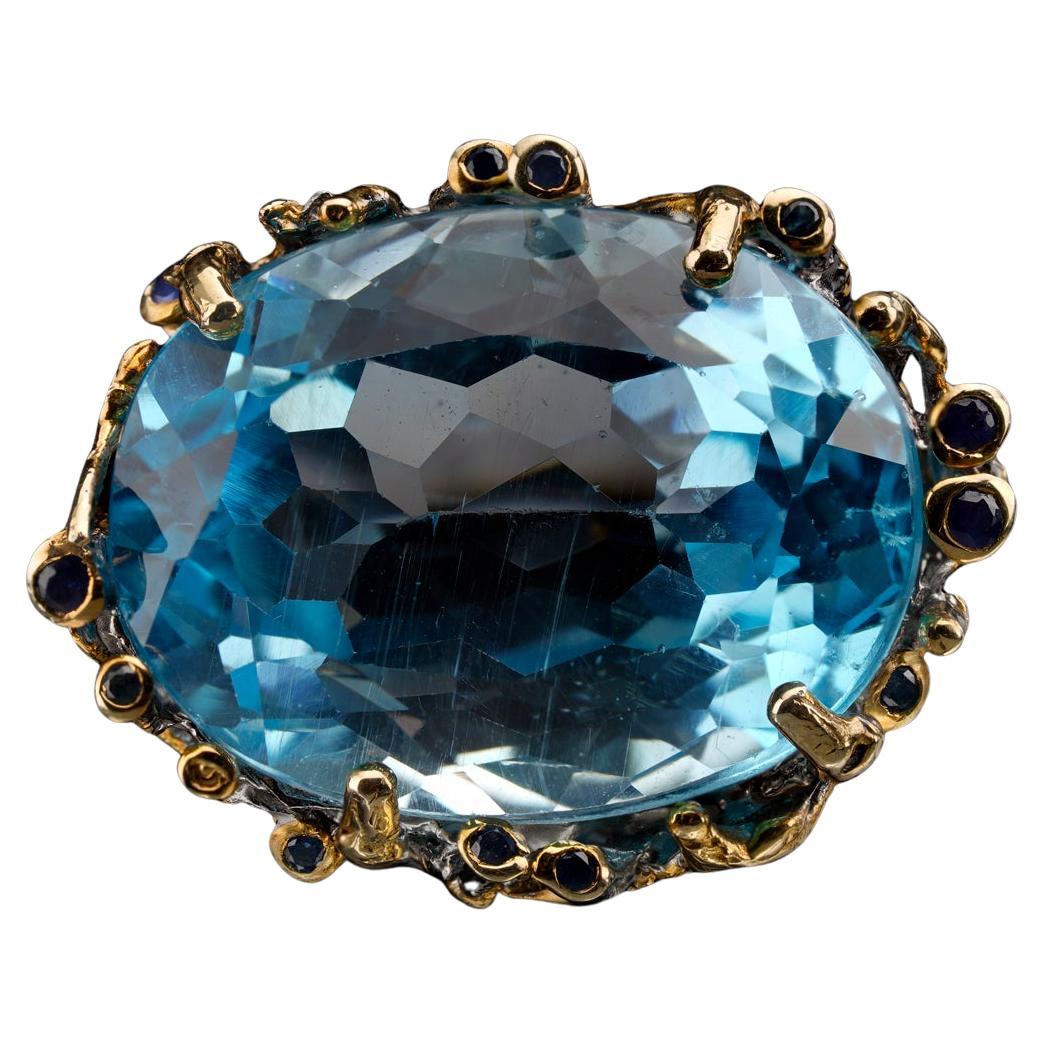 This massive, flawless oval-cut blue topaz fills a fantastically unique hand-tooled prong setting featuring carved bamboo shoots and accenting deep blue sapphires. A statement piece if ever there was one, this sterling silver ring features gold leaf