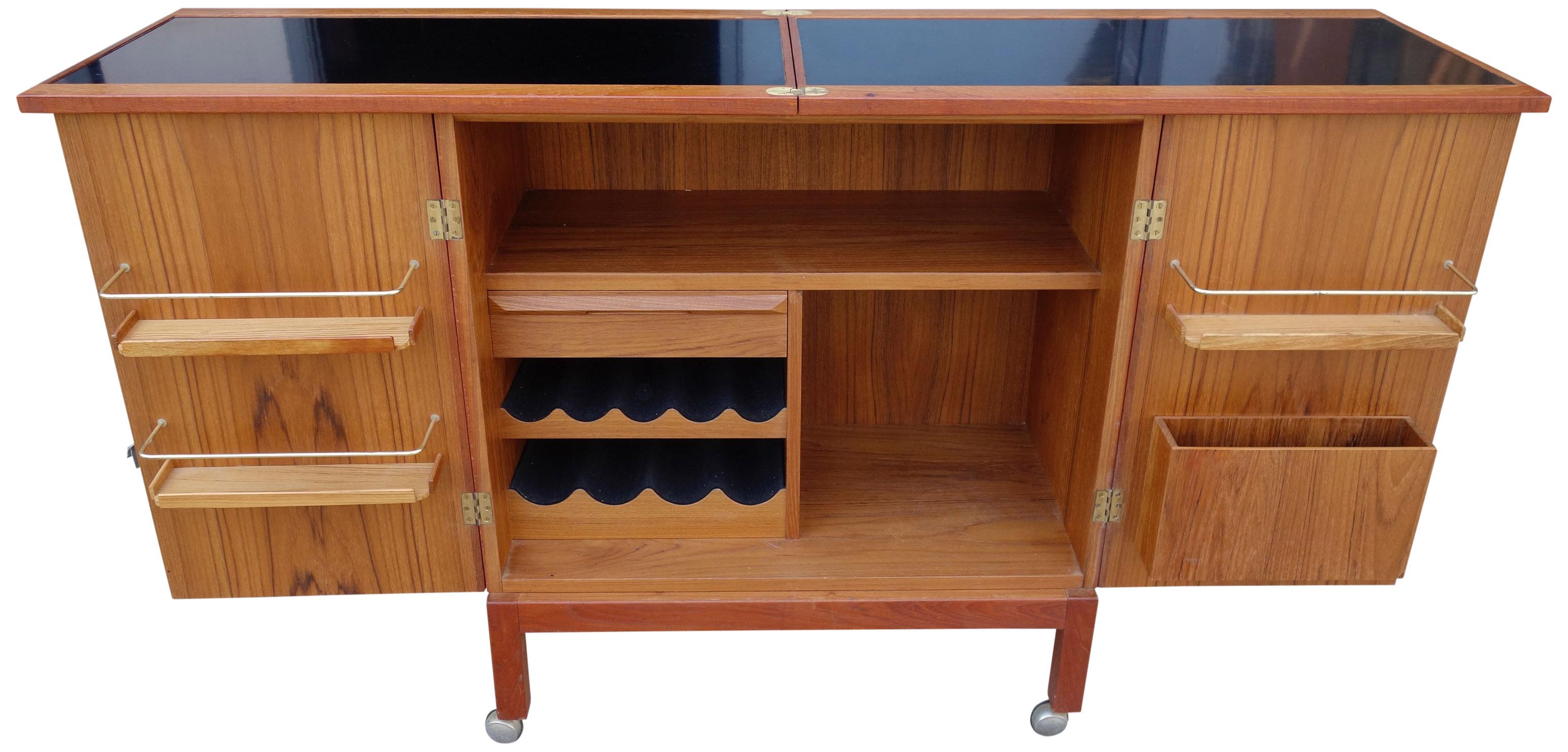 Scandinavian Modern at its best. Deceptively sleek and minimal on the outside but when opened and top extends revealing an amazingly crafted bar. This rolling teak bar cabinet features internal dovetailed drawers, wine storage and shelves with brass