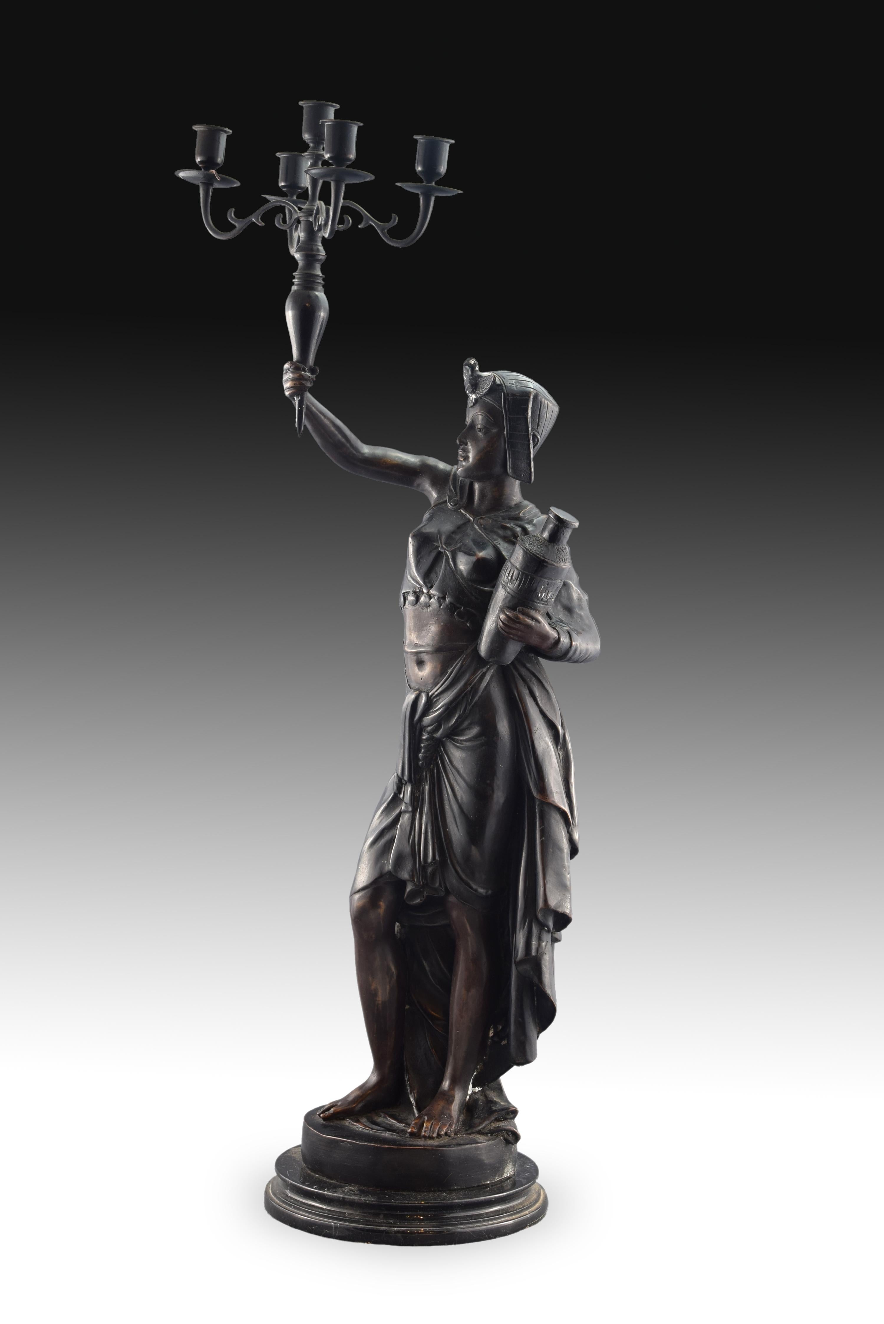 On a square base stands the figure of the woman, dressed according to what the19th century considered as Egyptian fashion. He holds a small pitcher with his left hand while raising with the right a torch that ends in a five-light candelabra. For