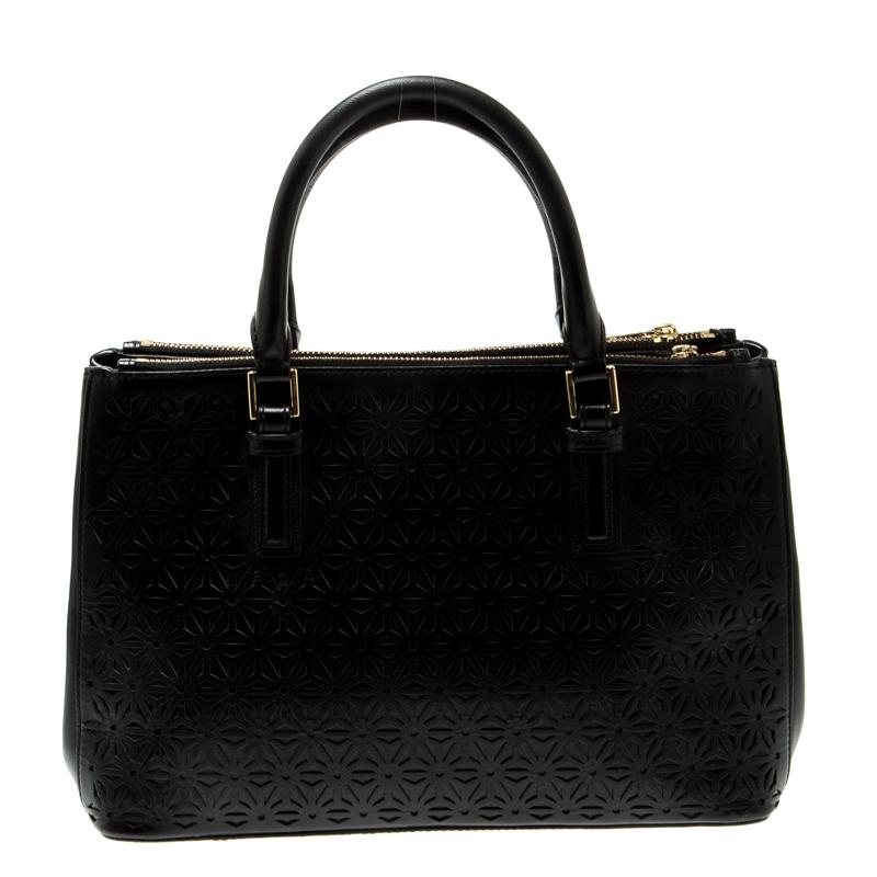 This stunning black tote is by Tory Burch. Crafted from leather, and lined with fabric on the insides, the bag features a lovely exterior adorned with floral laser cut design all over. It has dual rolled top handles, logo on the front, and two