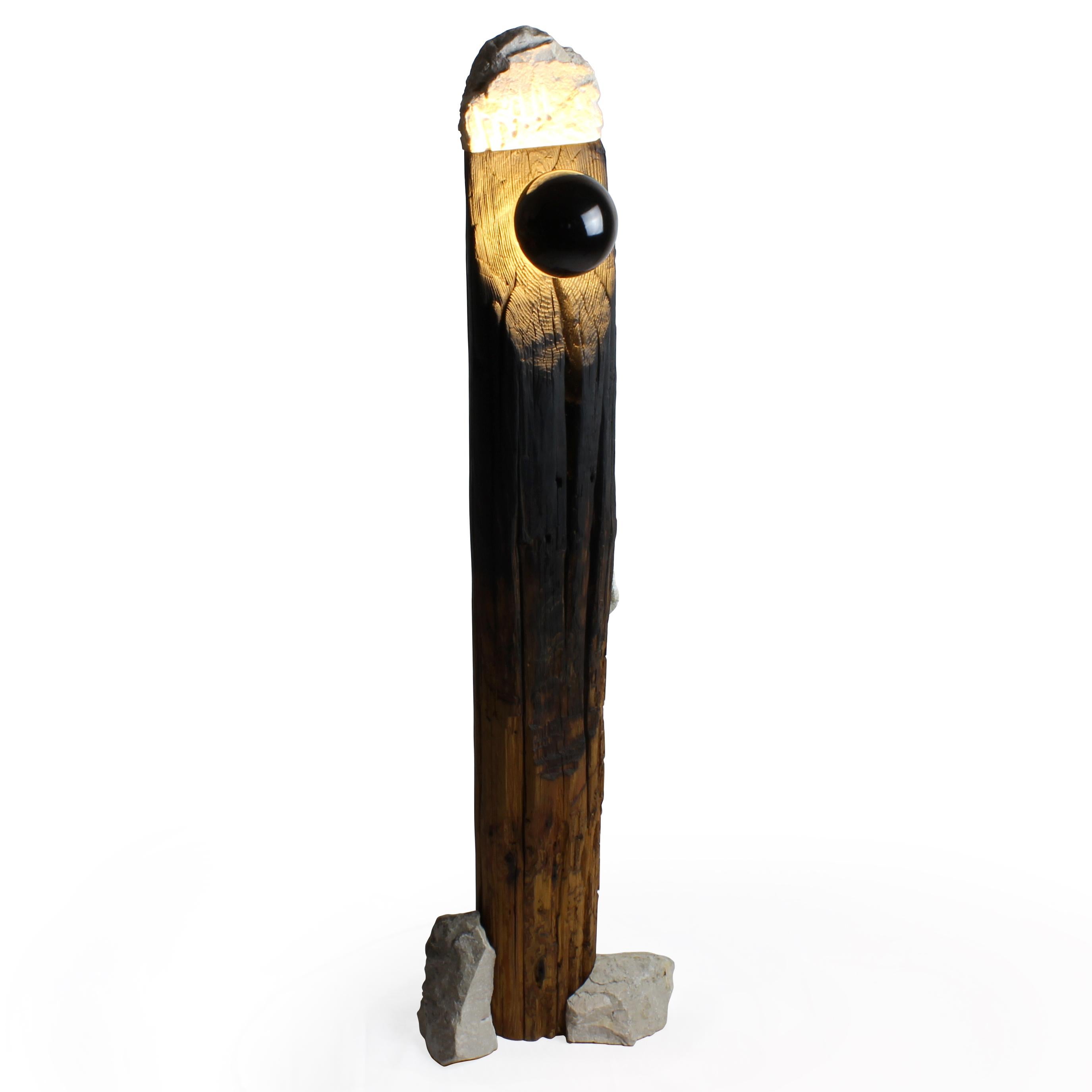 THIS DISTINCTIVE FLOOR LAMP EMANATES FROM AN ARTFULLY REPURPOSED AGED WOOD COLUMN,
COMPLEMENTED BY RESIDUAL STONE ELEMENTS. AS A SCULPTURAL ARTIFICE, ITS WOOD HAS BEEN
MASTERFULLY CHARRED USING THE YAKISUGI TECHNIQUE, WHILE THE STONE GRACING ITS